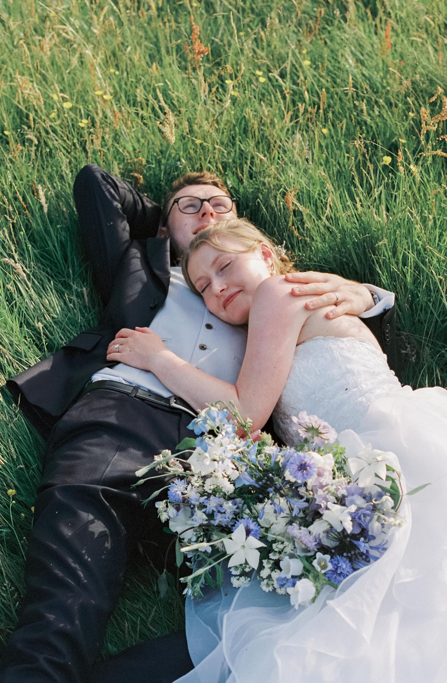 A couple lying in tall grass with a bouquet of blue and white flowers.