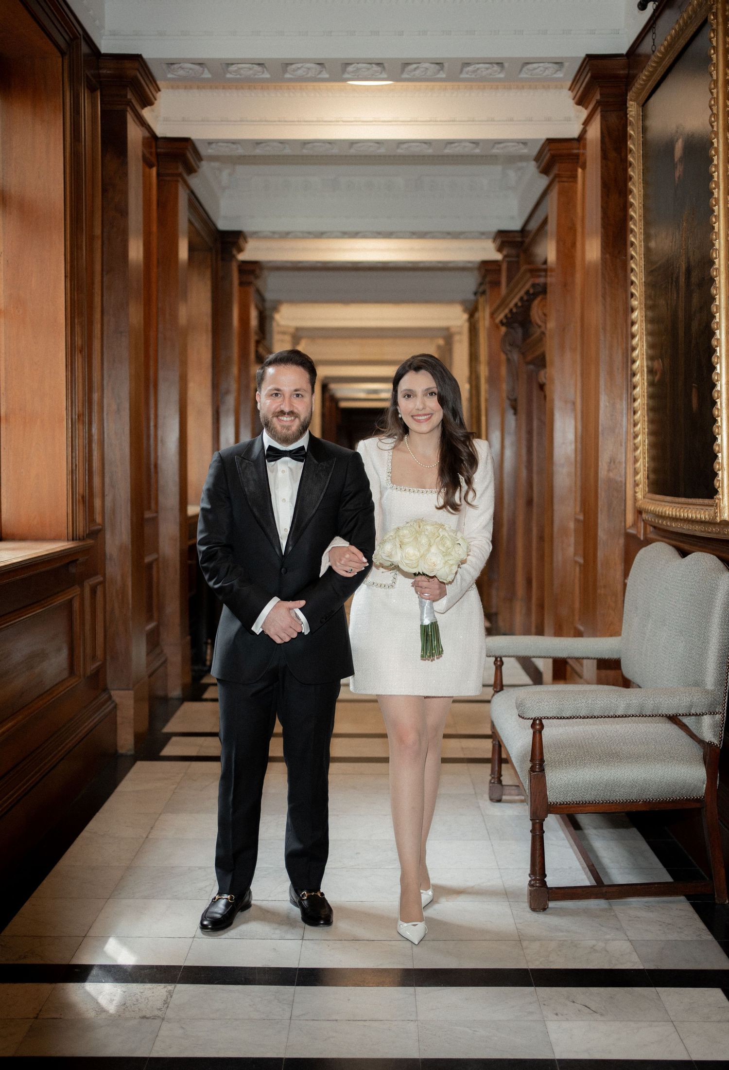 A couple poses playfully in formal attire against wooden paneled walls.