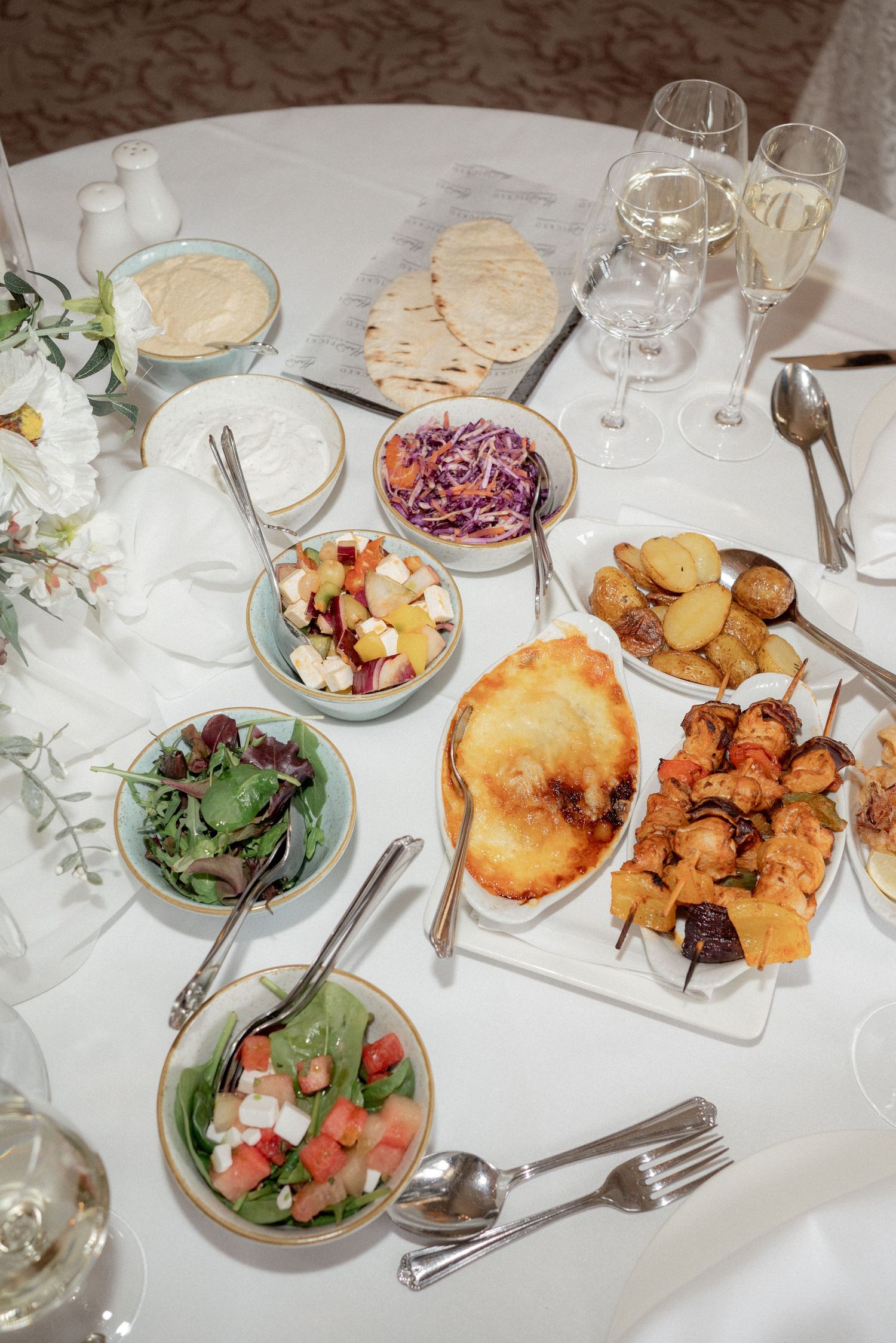Wedding reception table set with various appetizers and salads on white tablecloths.
