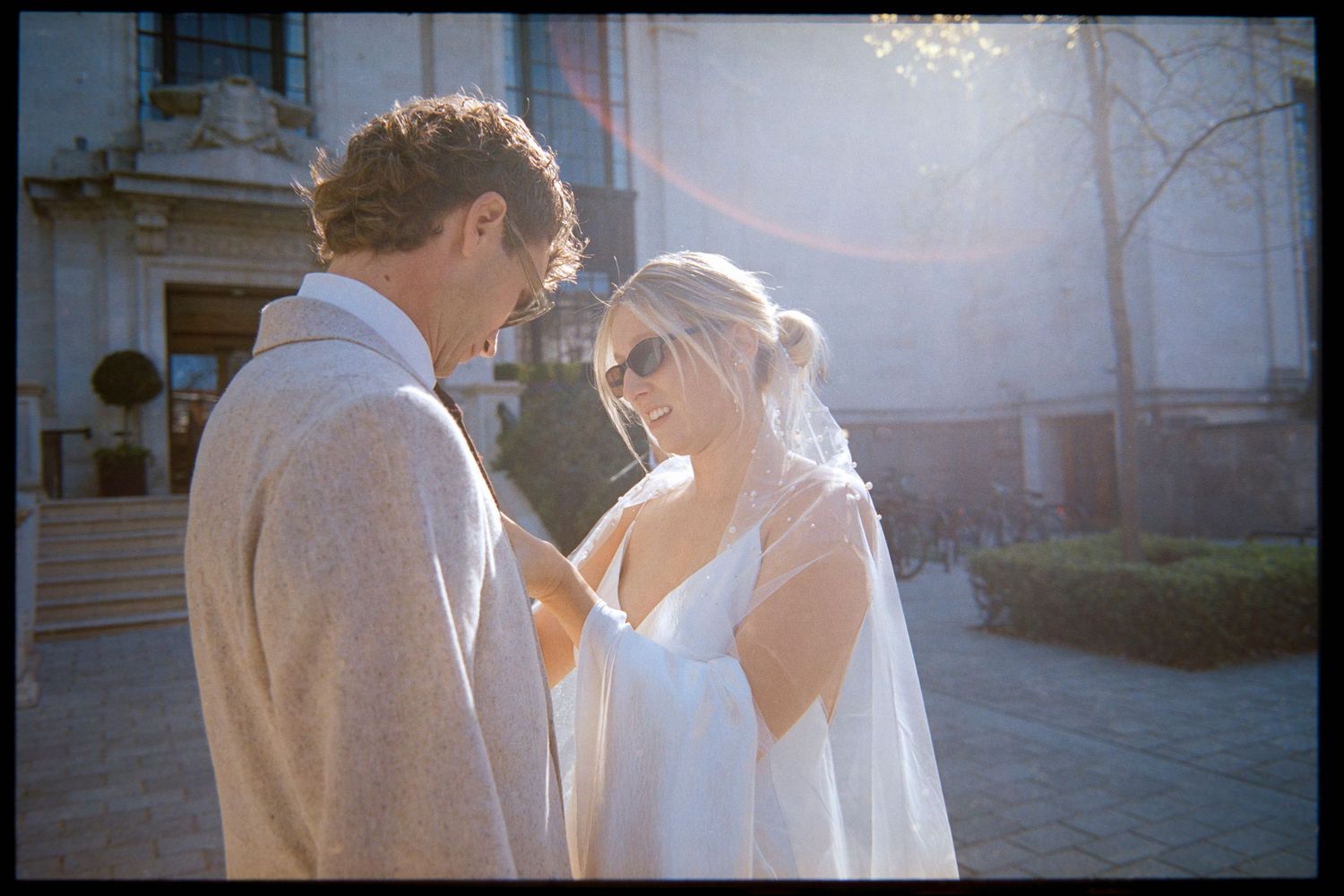 Bride in white dress and veil smiles at groom in gray suit outdoors in golden sunlight near elegant building.