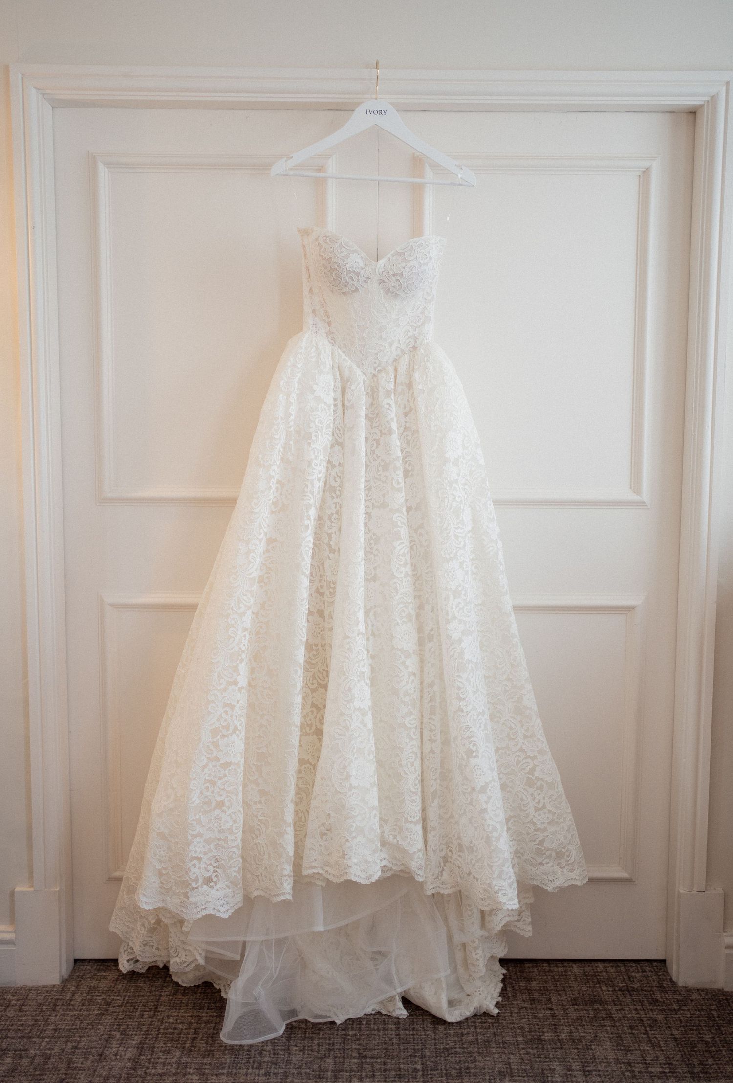White lace wedding dress hanging against paneled wall in preparation room.