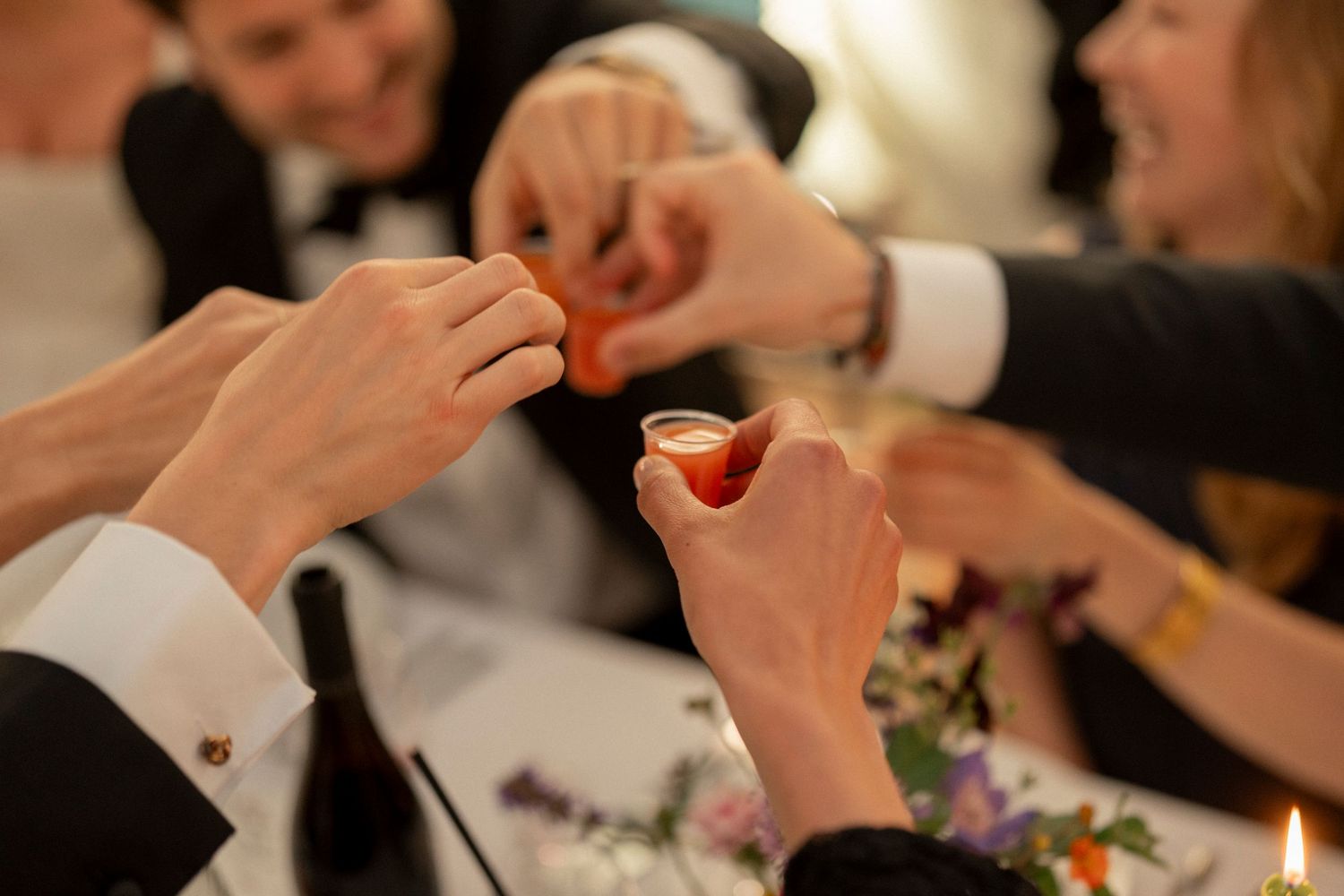 Guests at a wedding reception table raise small glasses in a toast, with hands clinking together beside a floral centerpiece and candle.