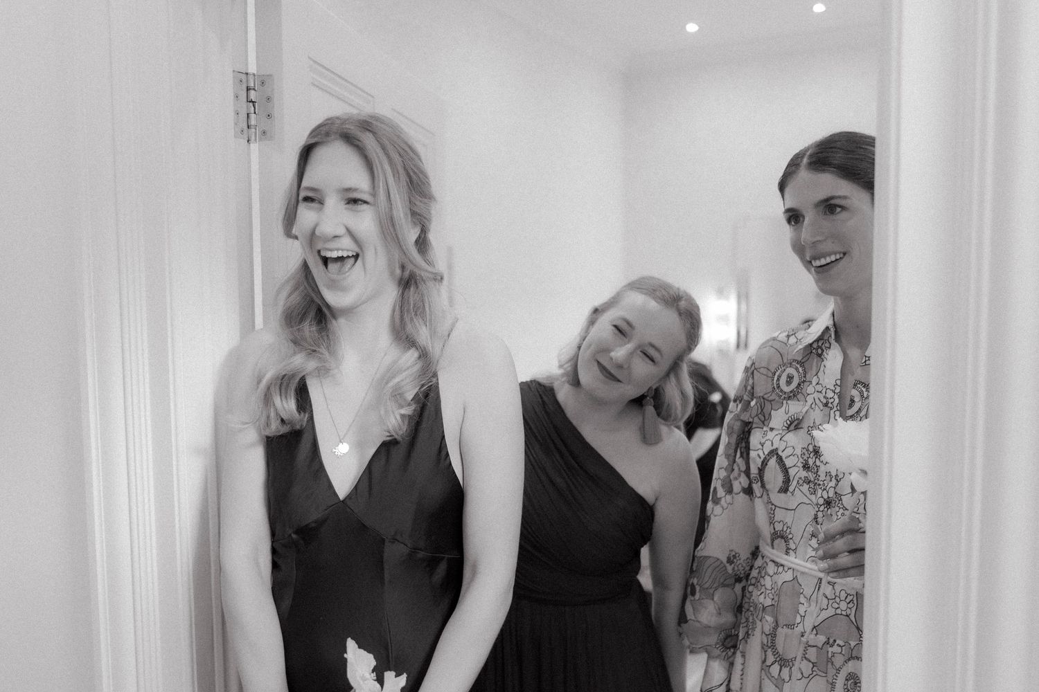 Three women in elegant dresses laughing together in a bright doorway, captured in a joyful black and white candid moment.