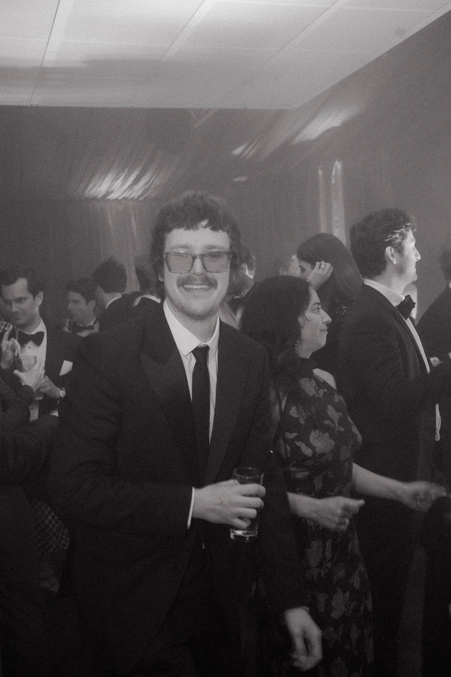 Black and white photo of a smiling man in glasses, mustache, and dark suit holding a drink at a crowded formal evening party.