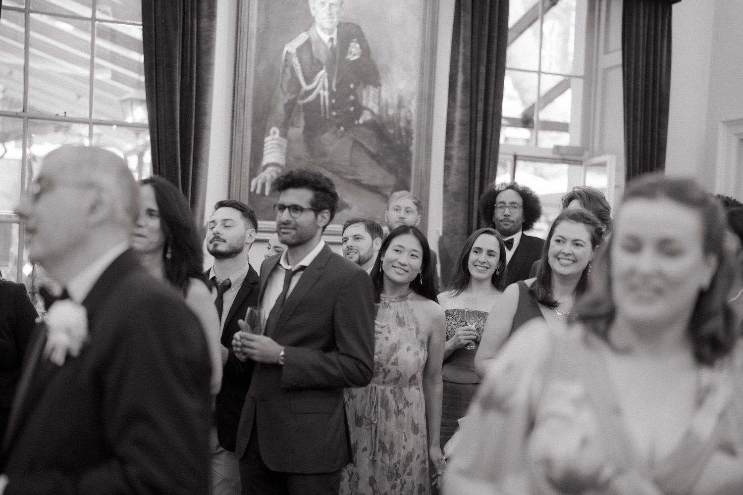 Black and white photo of wedding guests socializing beneath a portrait at elegant reception venue.
