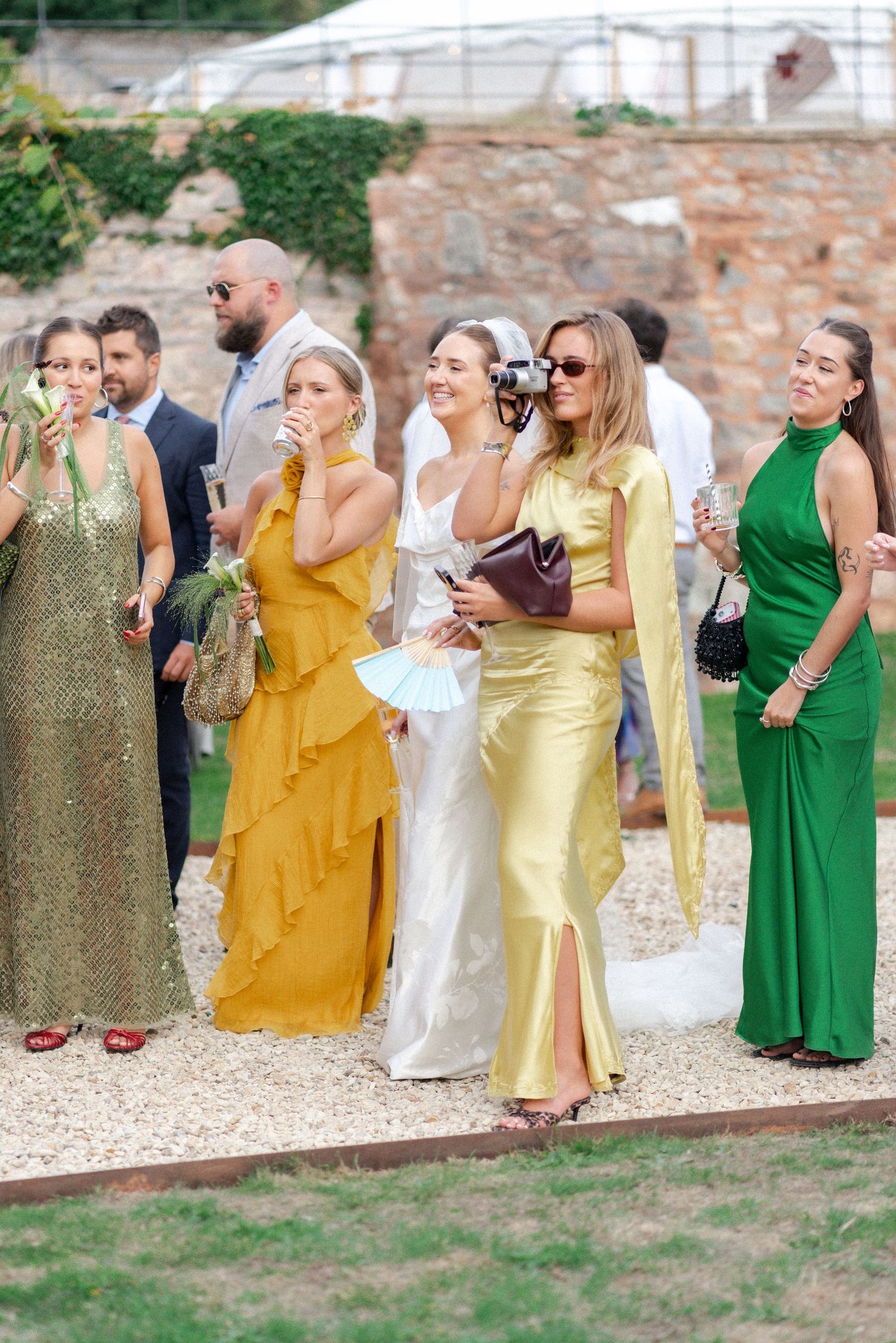 Wedding guests in vibrant yellow, green, and gold gowns gather outdoors at a sunny garden wedding celebration.