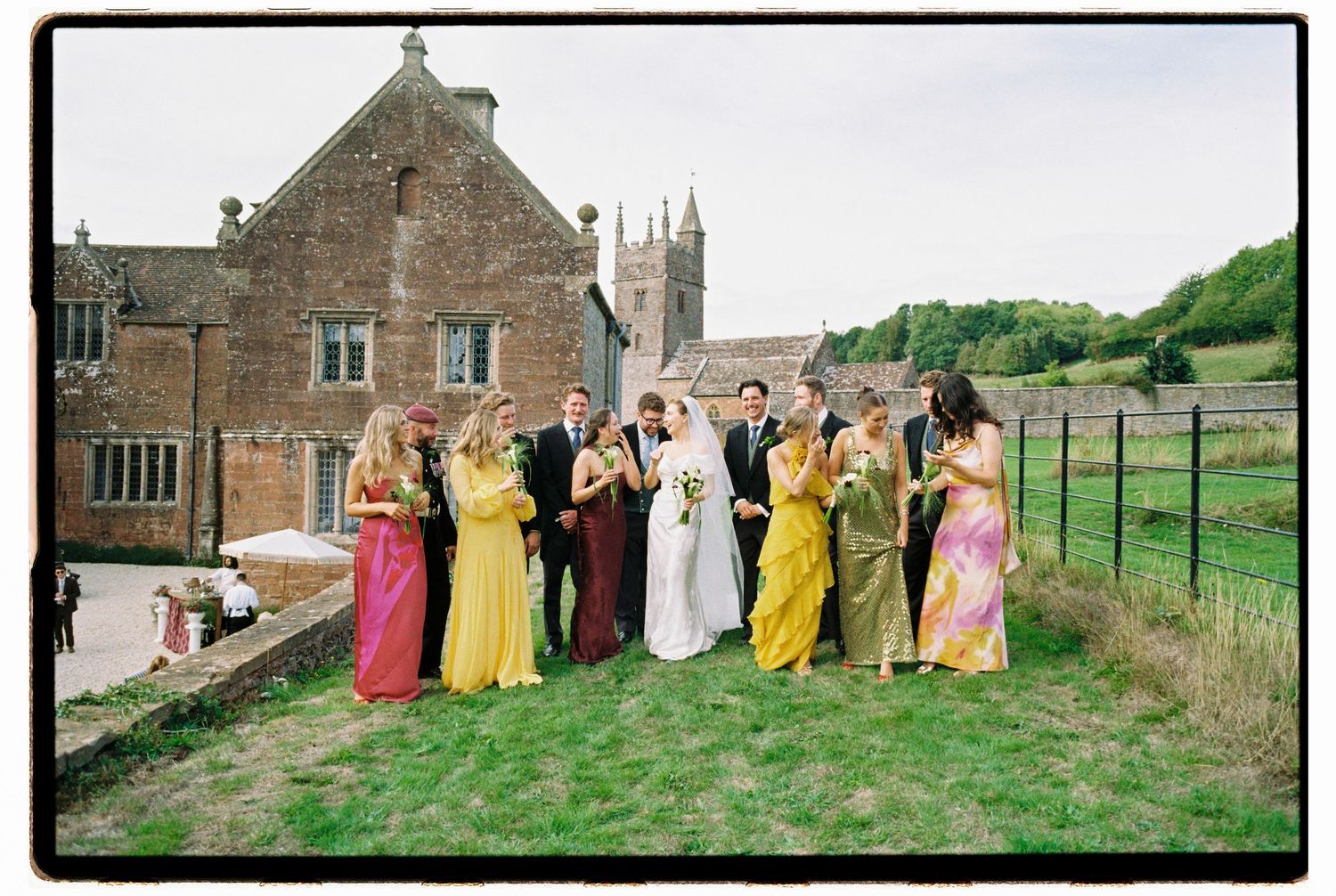 Wedding party in colorful bridesmaid dresses poses outside a historic stone manor church in the English countryside.