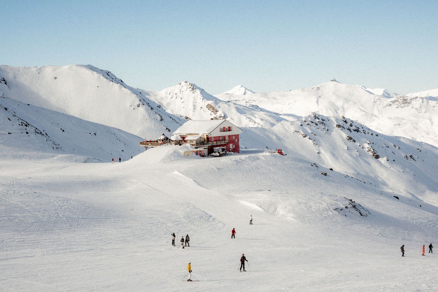 Skiers descend snowy slopes at a mountain resort with snow-capped peaks and blue sky in the background.