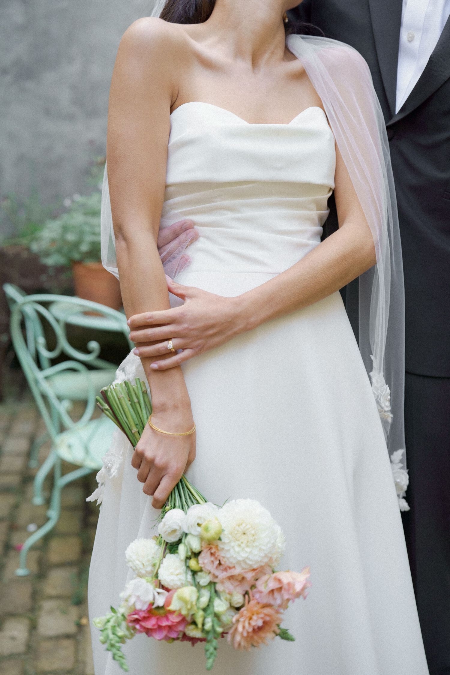 A romantic wedding couple embraces while holding a delicate bouquet of pink and white flowers against a stone wall.