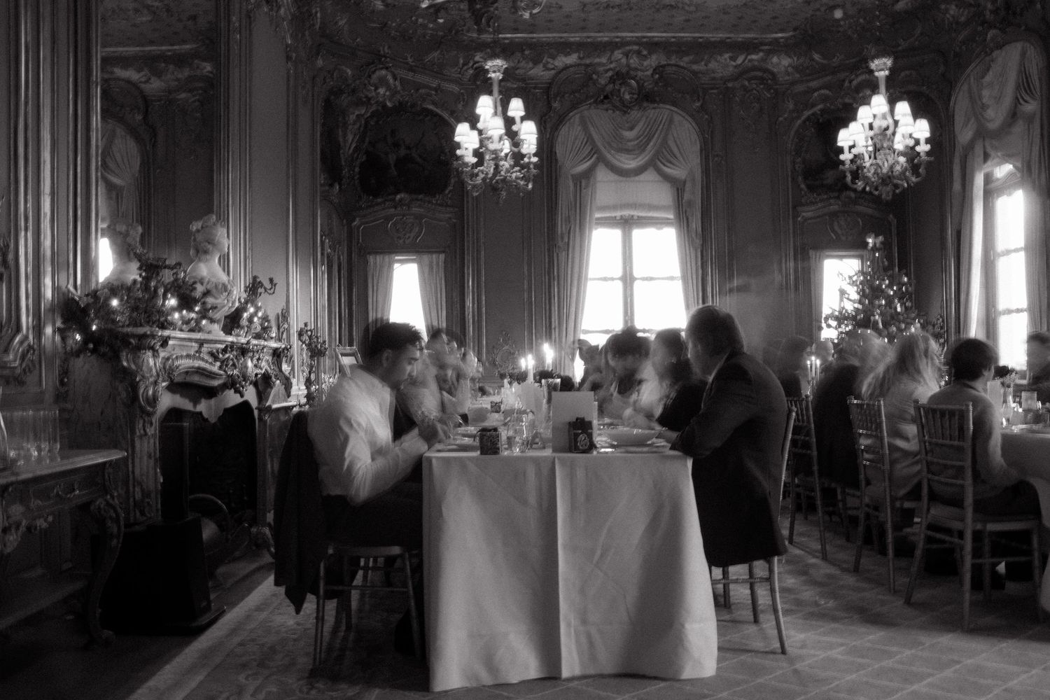 Elegant black and white photo of guests dining in an ornate chandelier-lit ballroom with candles.