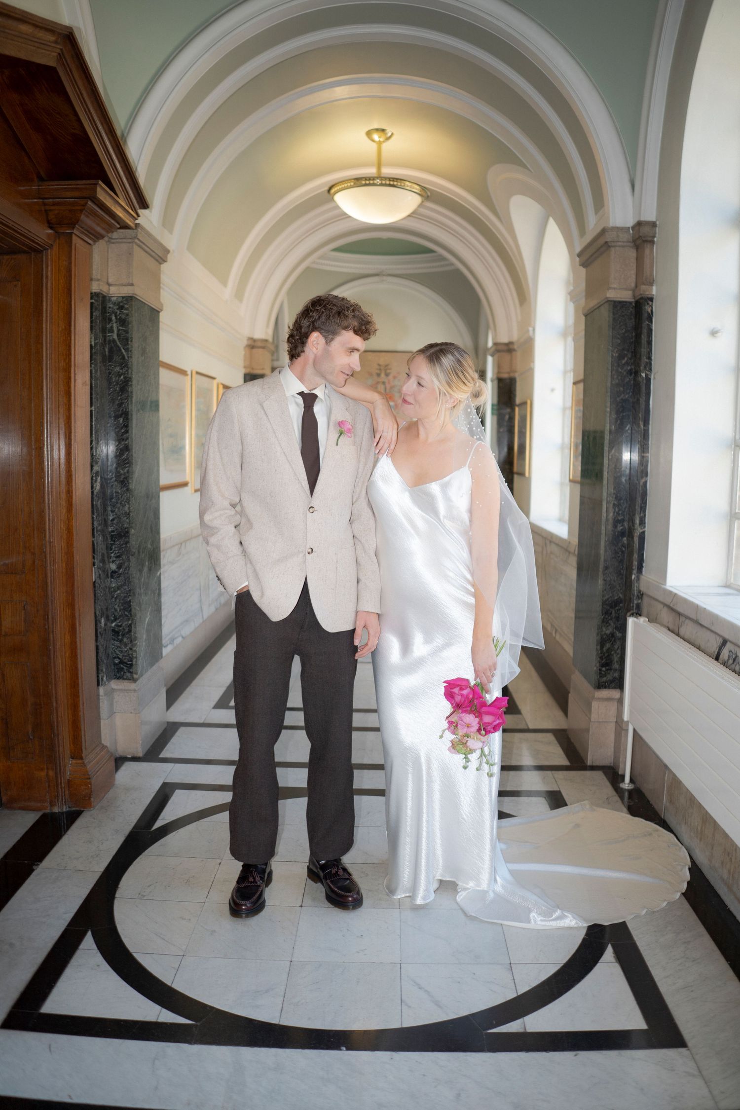 Bride and groom stand together in an elegant marble hallway, she holds pink flowers in a white satin gown.
