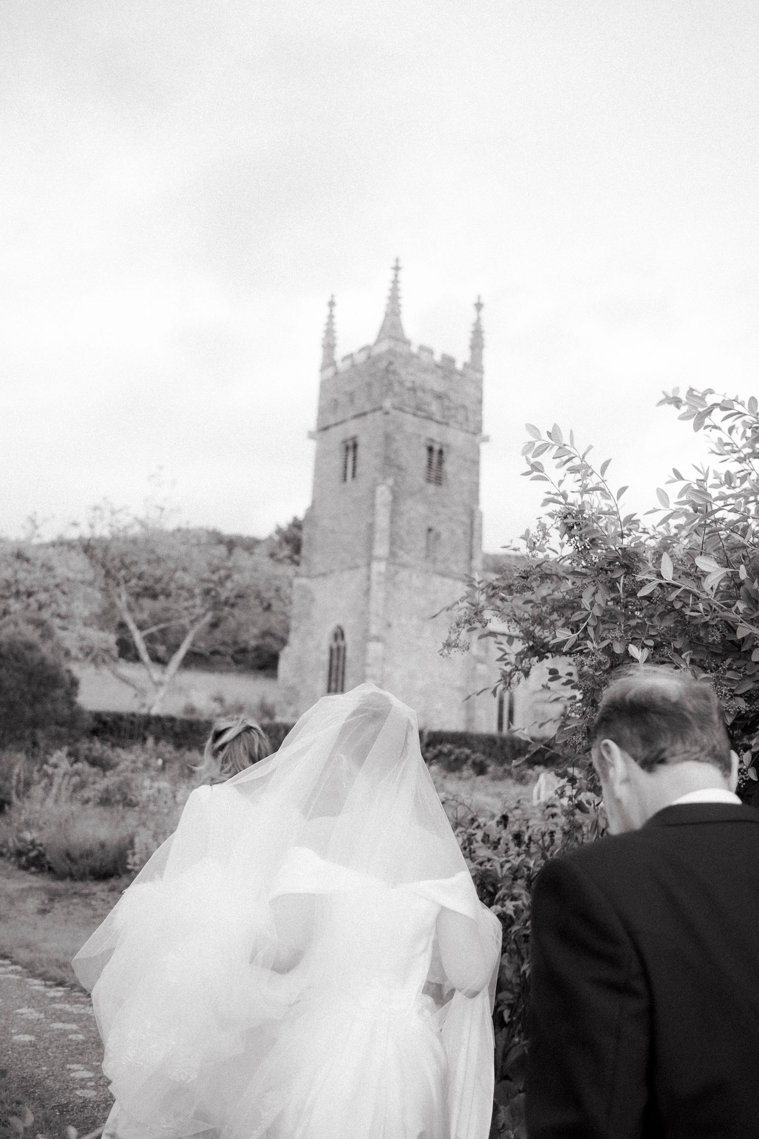 Bride in flowing veil walks toward historic stone church tower with groom in black and white wedding photo.
