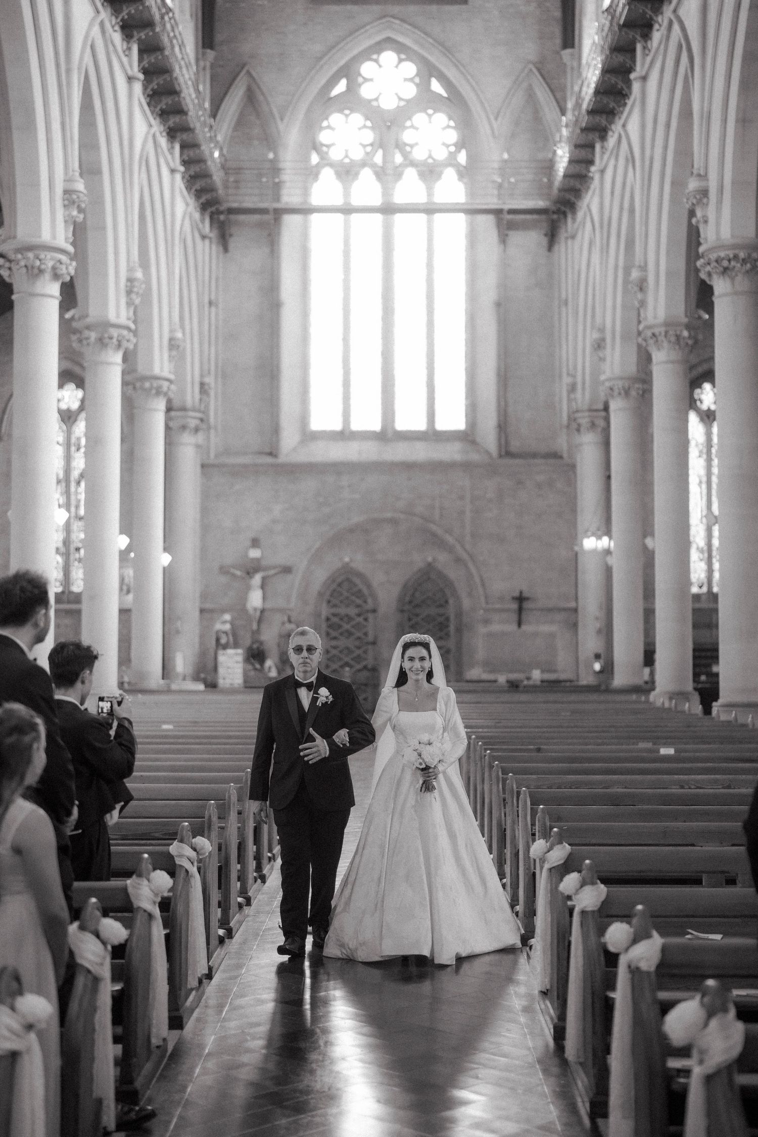 Black and white photo of a bride walking down the aisle with her father in a grand Gothic church.