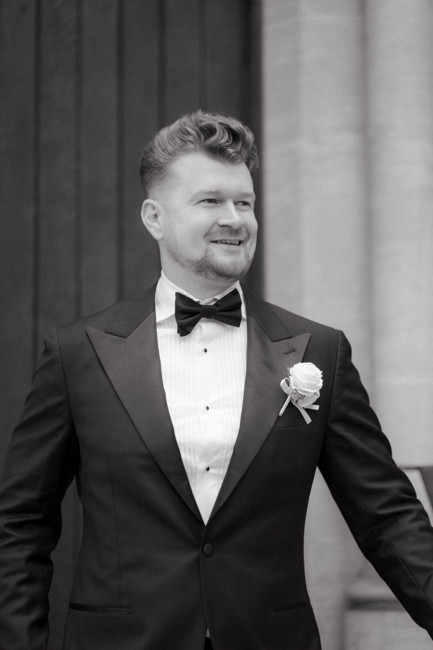 Elegant groom in black tuxedo with bow tie and white boutonniere smiling at outdoor wedding ceremony.