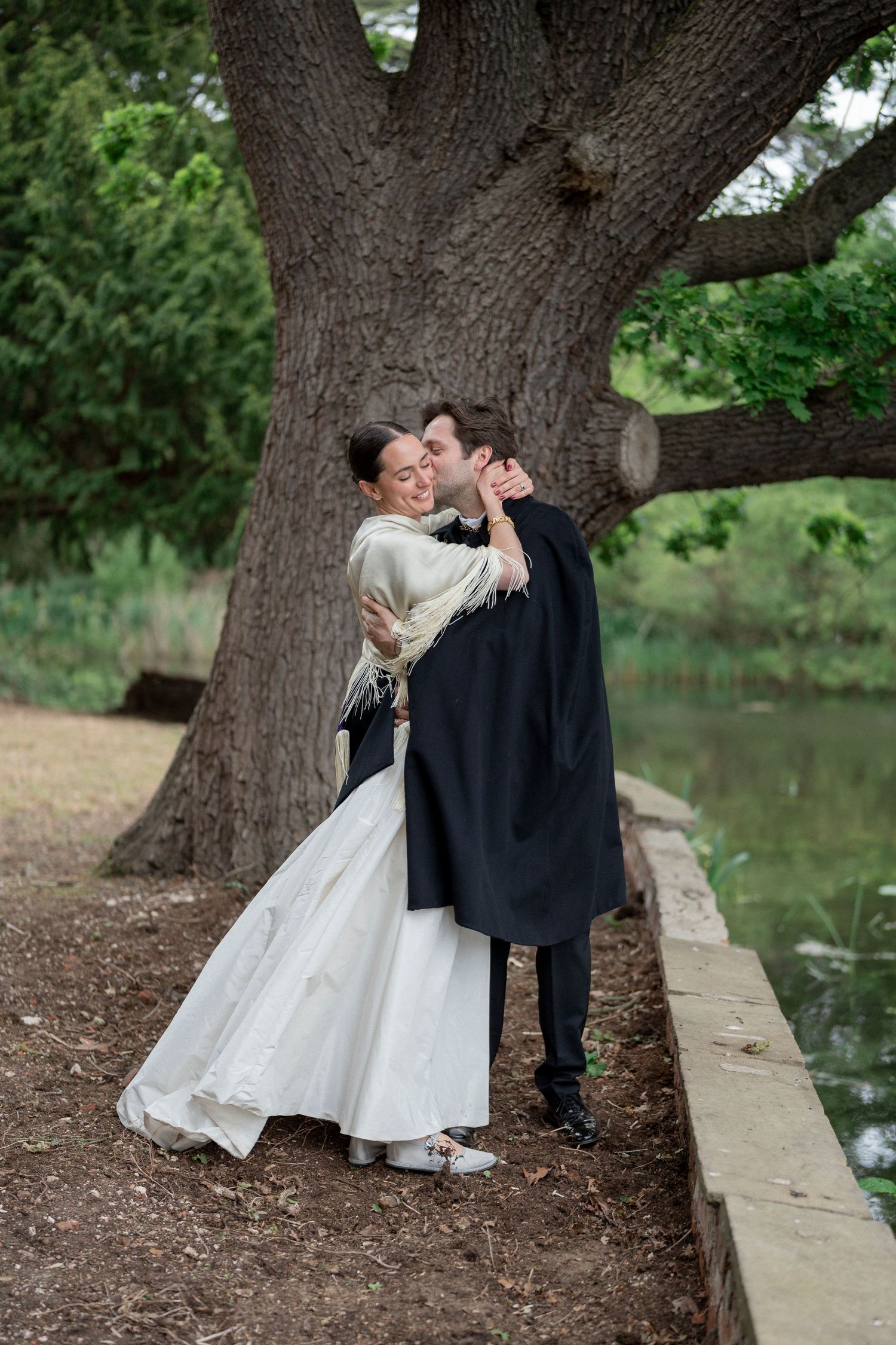 A bride and groom embrace and kiss beneath a large old tree beside a calm pond at their outdoor wedding.