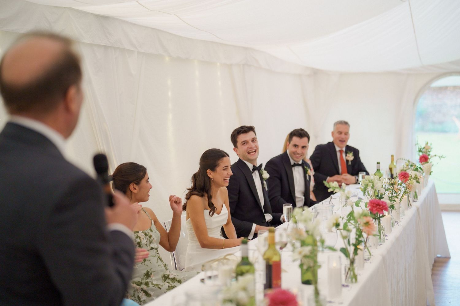 Wedding guests seated at long tables decorated with floral centerpieces enjoy dinner and conversation at an elegant reception.