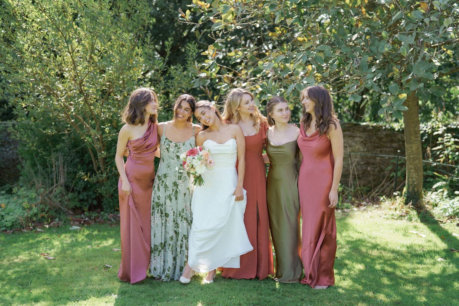 Bridesmaids in dusty rose dresses and a bride in white pose together in a lush garden setting on a sunny day.
