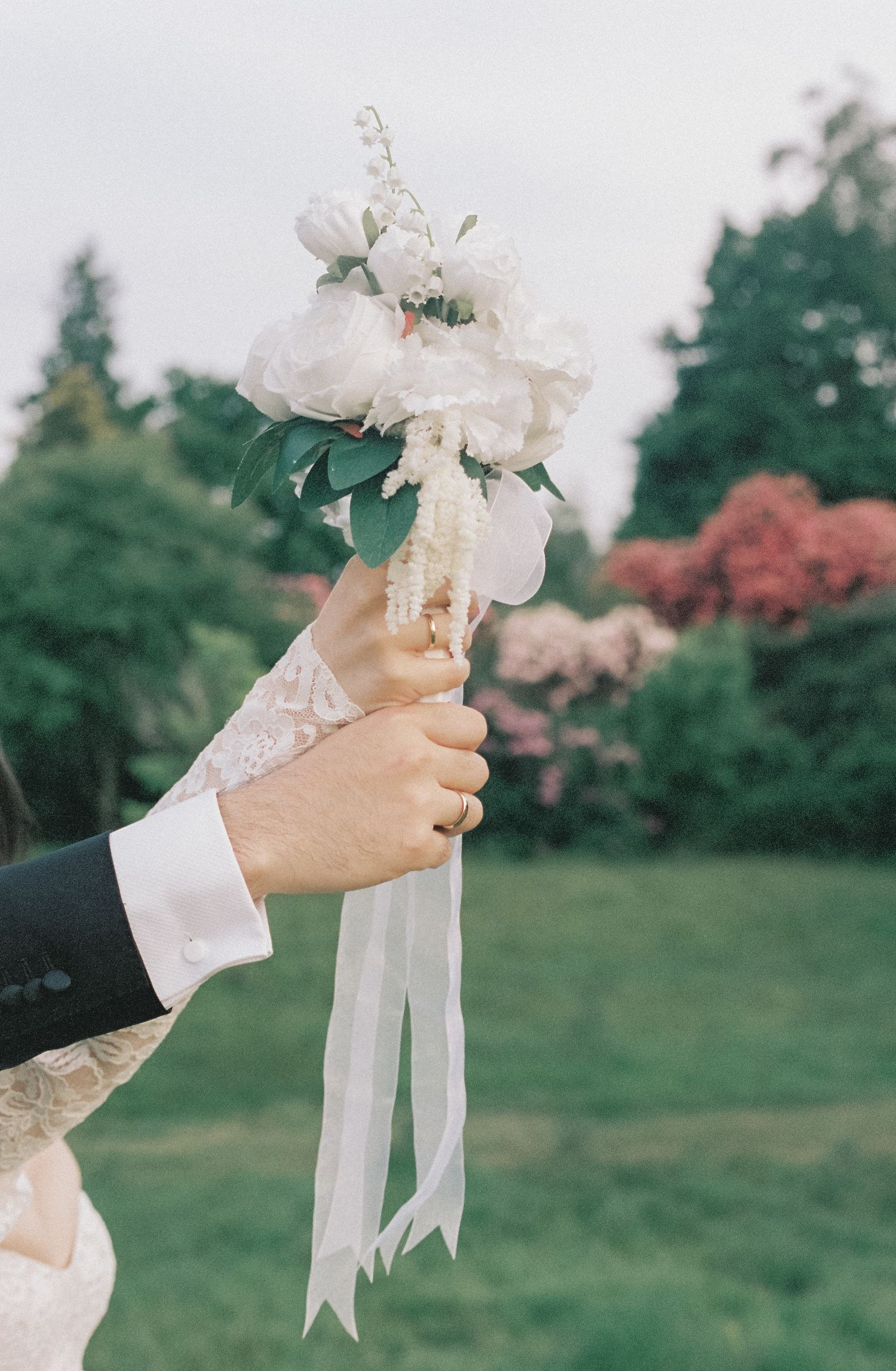 Hand holding a delicate white wedding bouquet with flowing ribbons against a garden background.