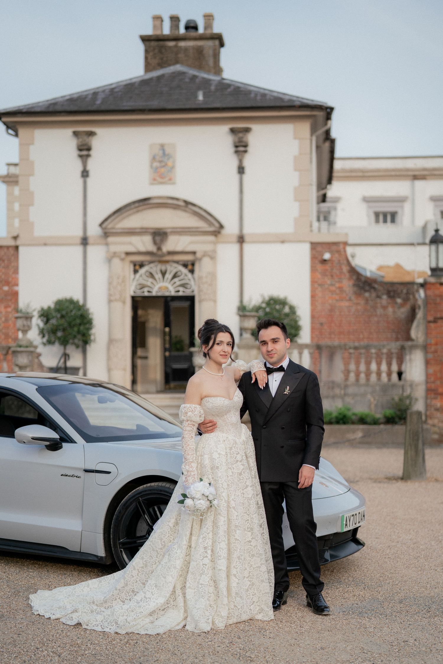 A wedding couple poses outside a grand historic building with their luxury car.