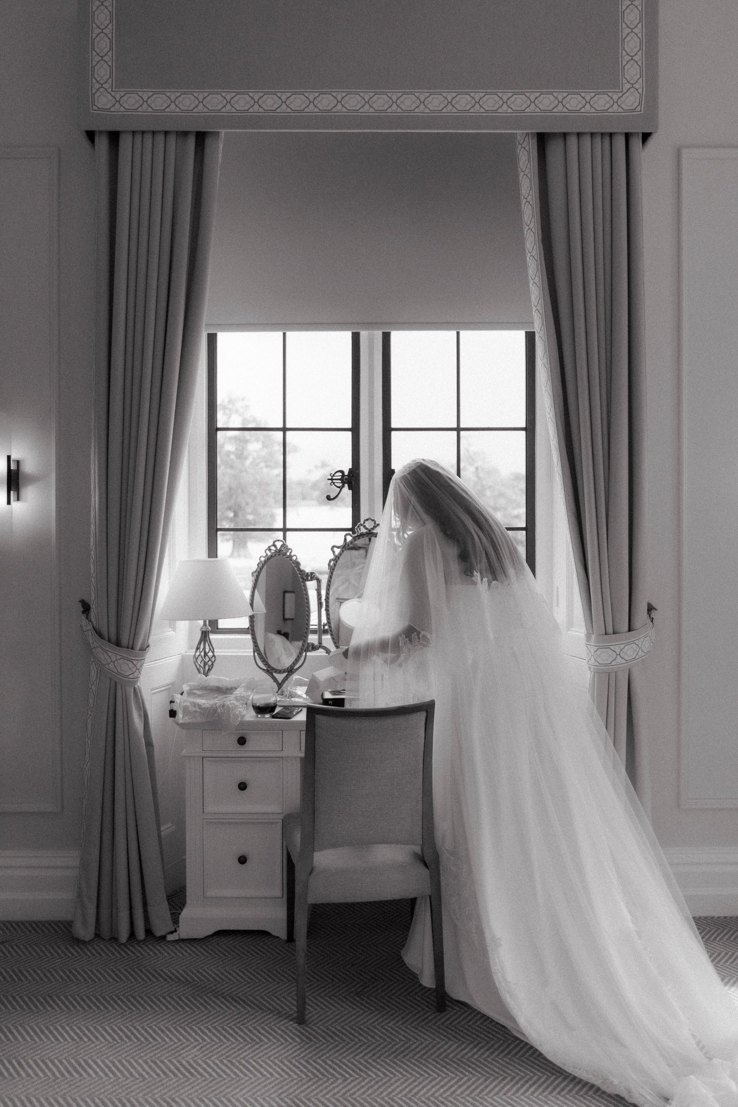 Bride in flowing wedding gown and veil sits at vintage dressing table by window in black and white photo.