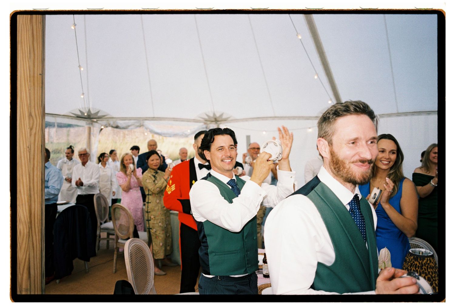Two grooms in green vests clapping and smiling during wedding reception celebration inside marquee tent.