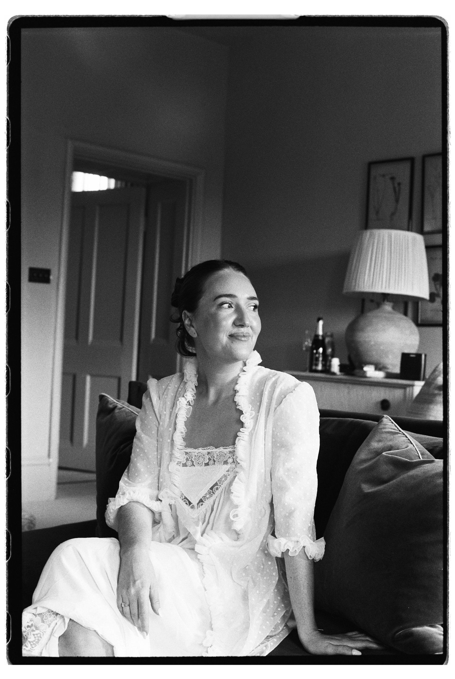 Black and white photo of a woman in a white dress sitting on a sofa, gazing thoughtfully in an elegant room.