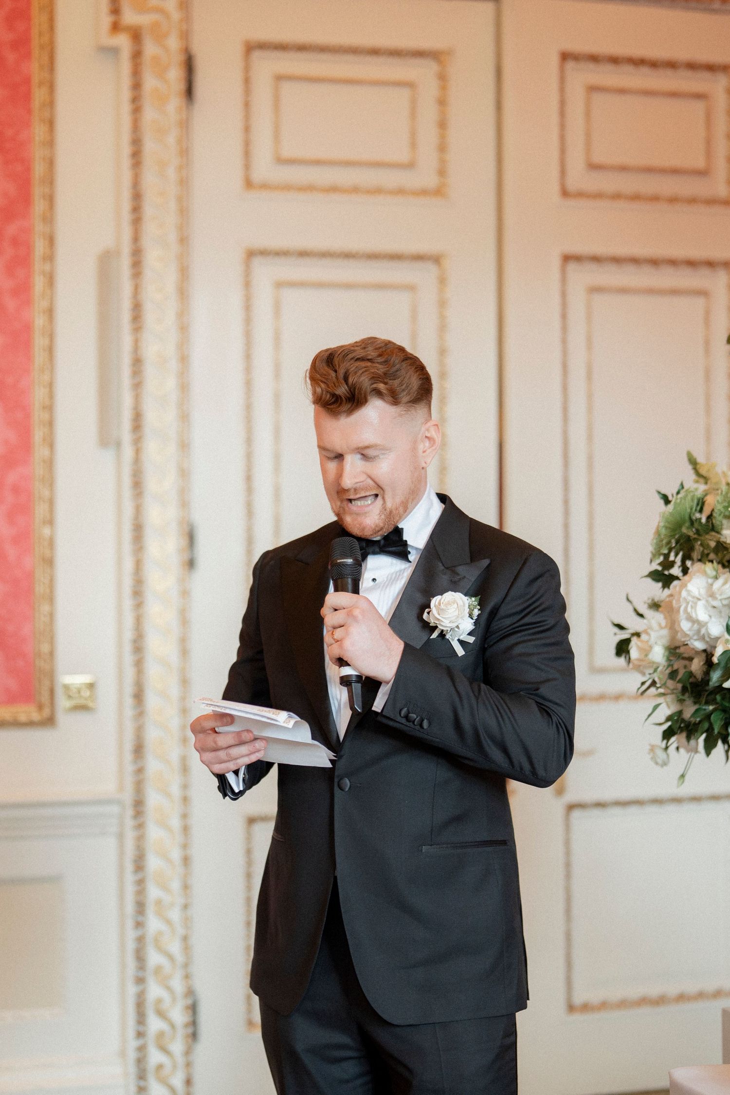 Groom in black tuxedo gives speech holding microphone and notes at wedding reception.