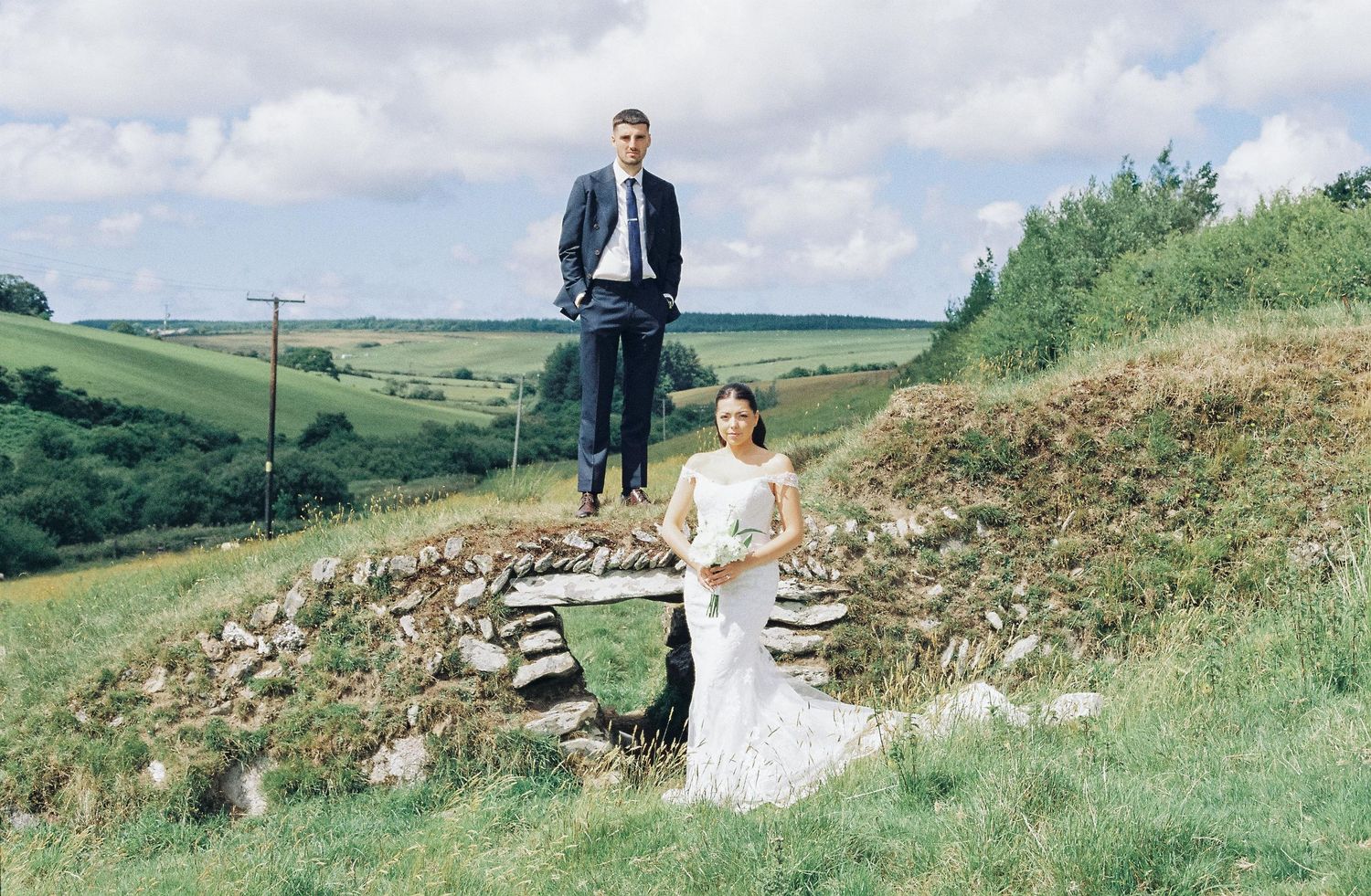 Panoramic landscape view of a wedding couple standing on a hillside against cloudy skies.
