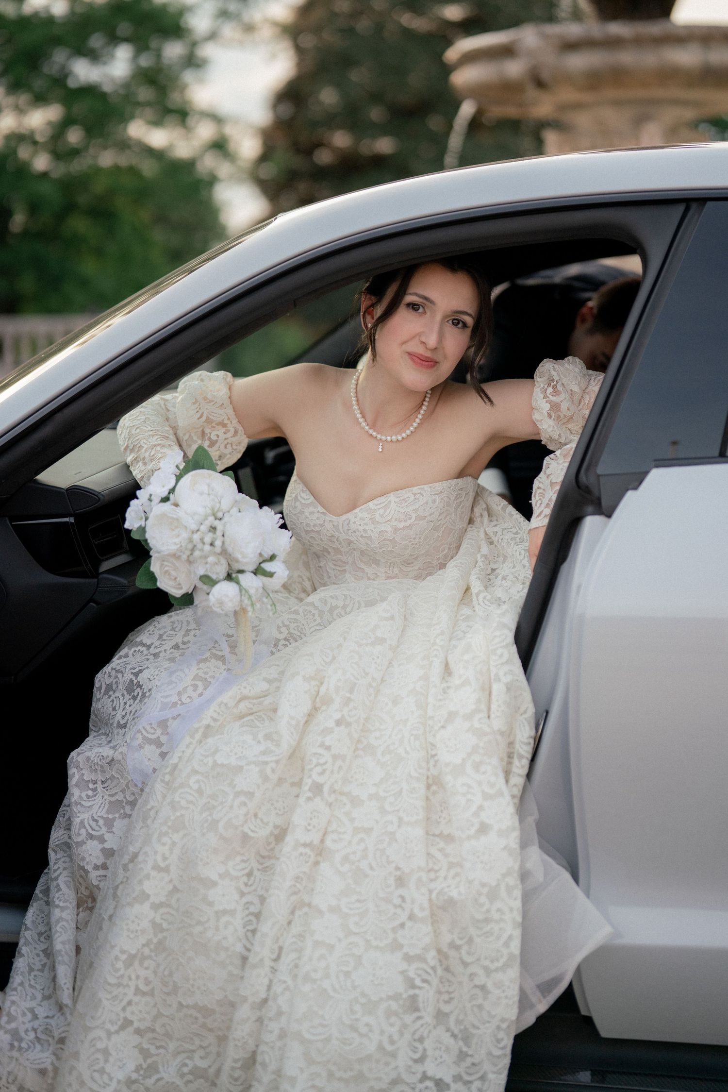 An elegant bride in a lace wedding dress sits in a white car holding a bouquet.
