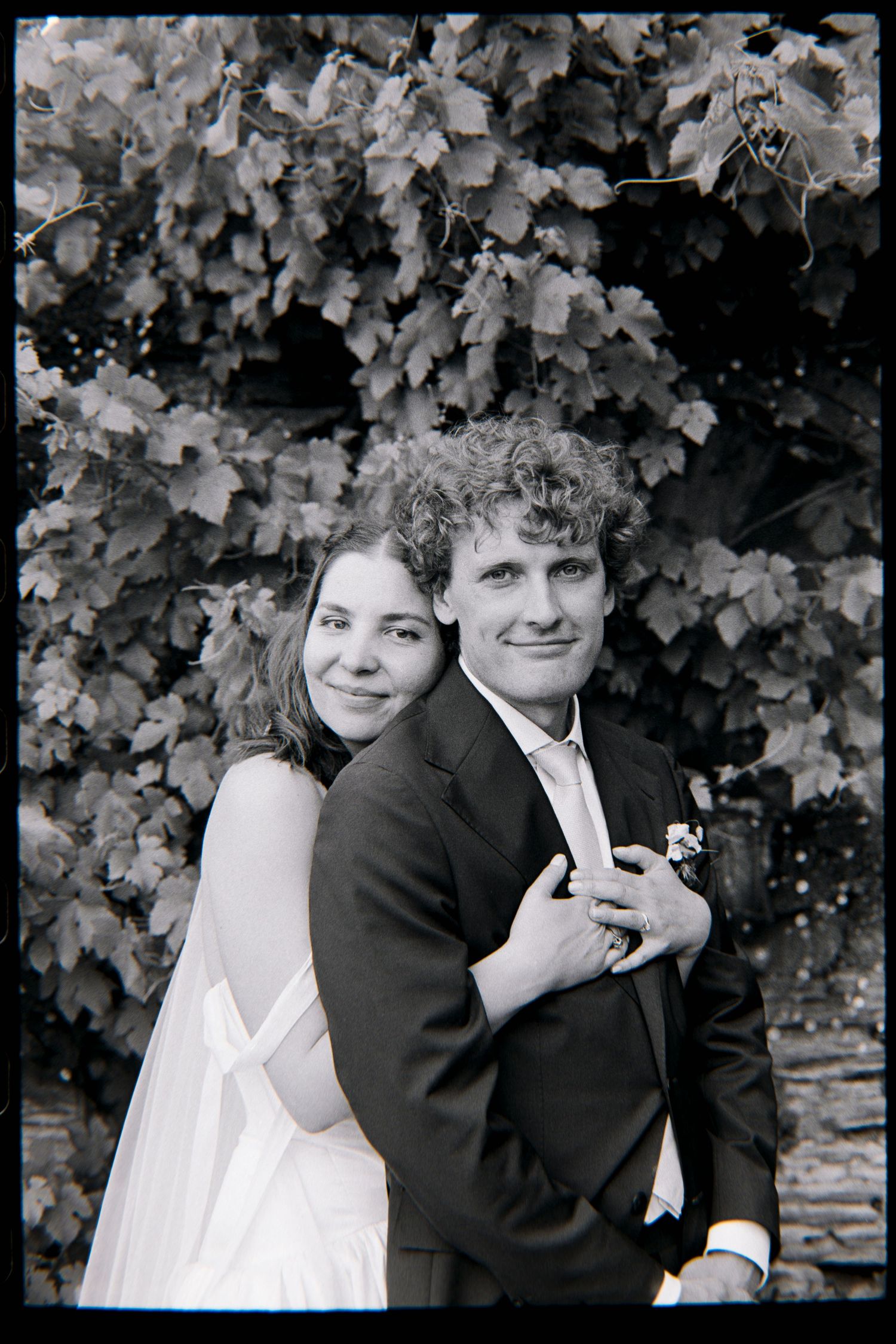 Black and white wedding photo of a bride in white dress embracing groom in dark suit before leafy backdrop.