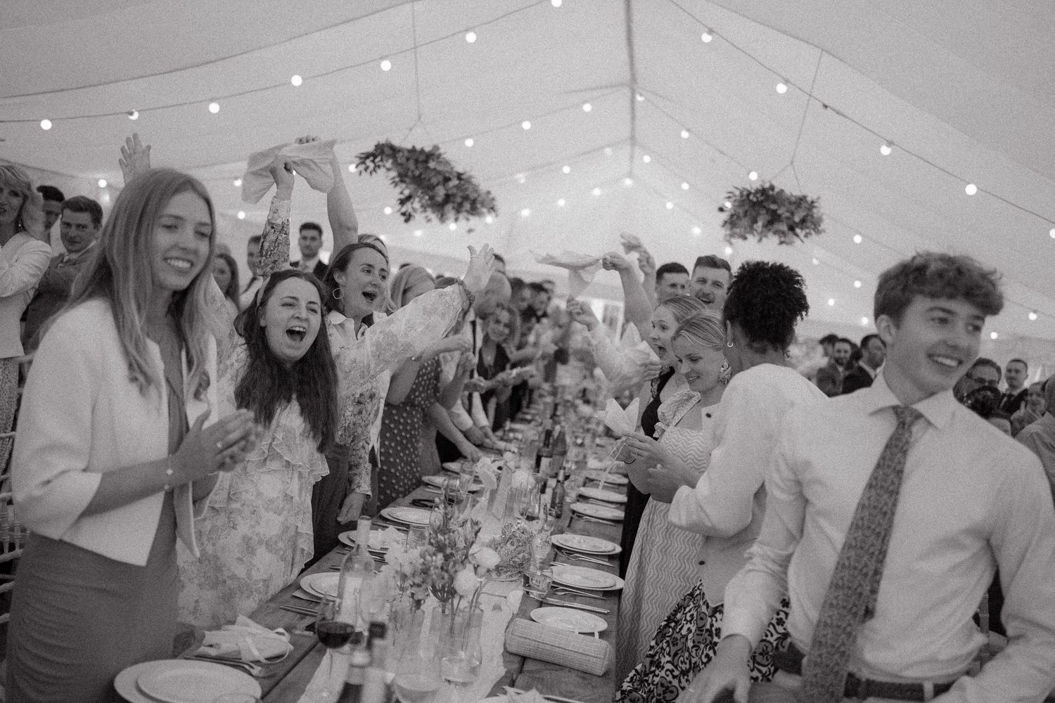 Black and white photo of wedding reception guests standing and celebrating inside a tent with string lights overhead.