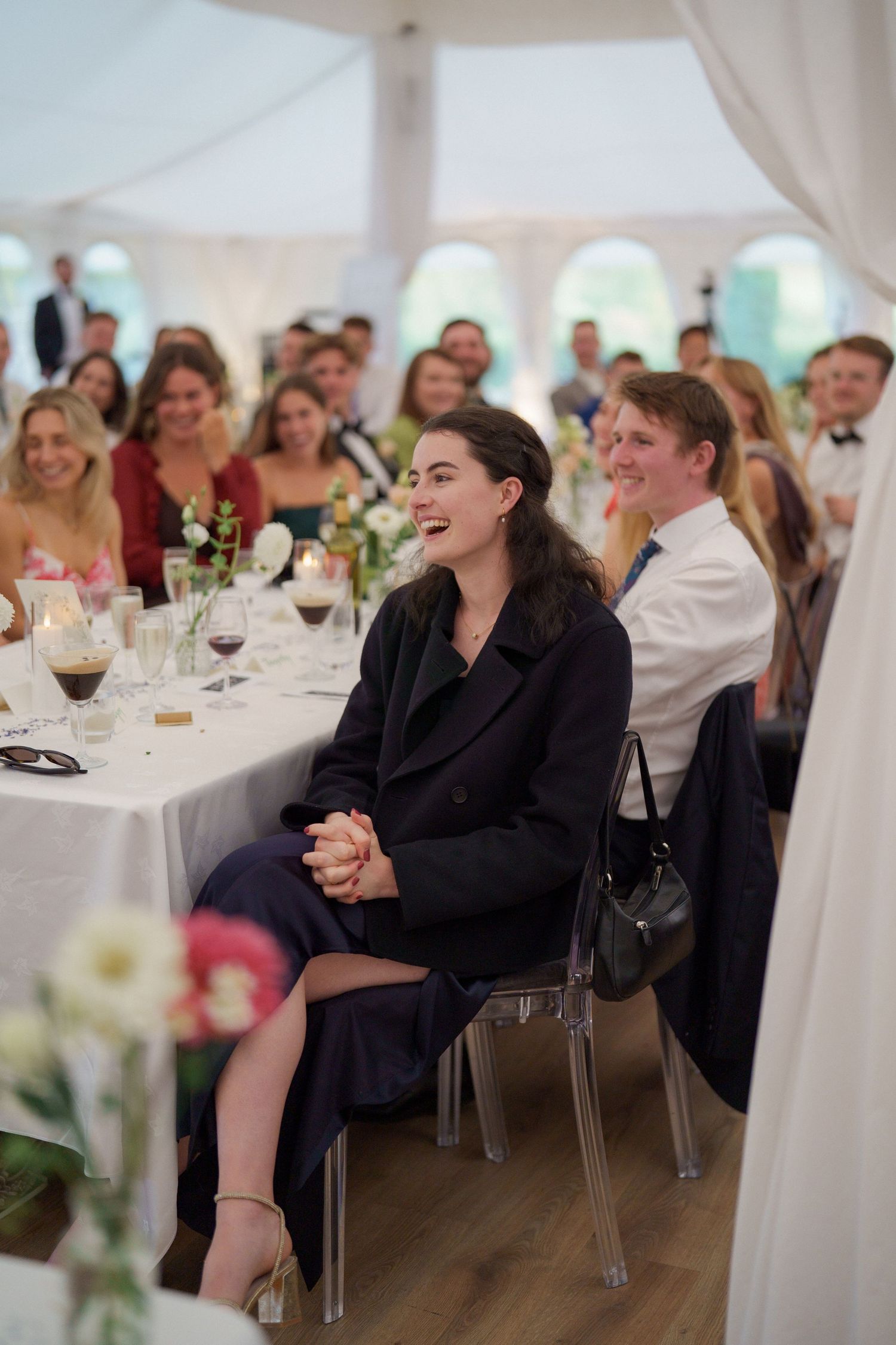 Wedding guests laugh and enjoy themselves during a reception under an elegant white tent with table settings and flowers.