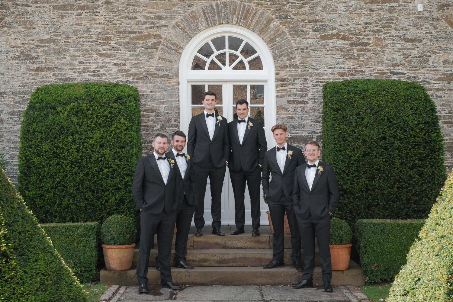 Groomsmen in black tuxedos stand on stone steps between manicured hedges at an elegant estate entrance.