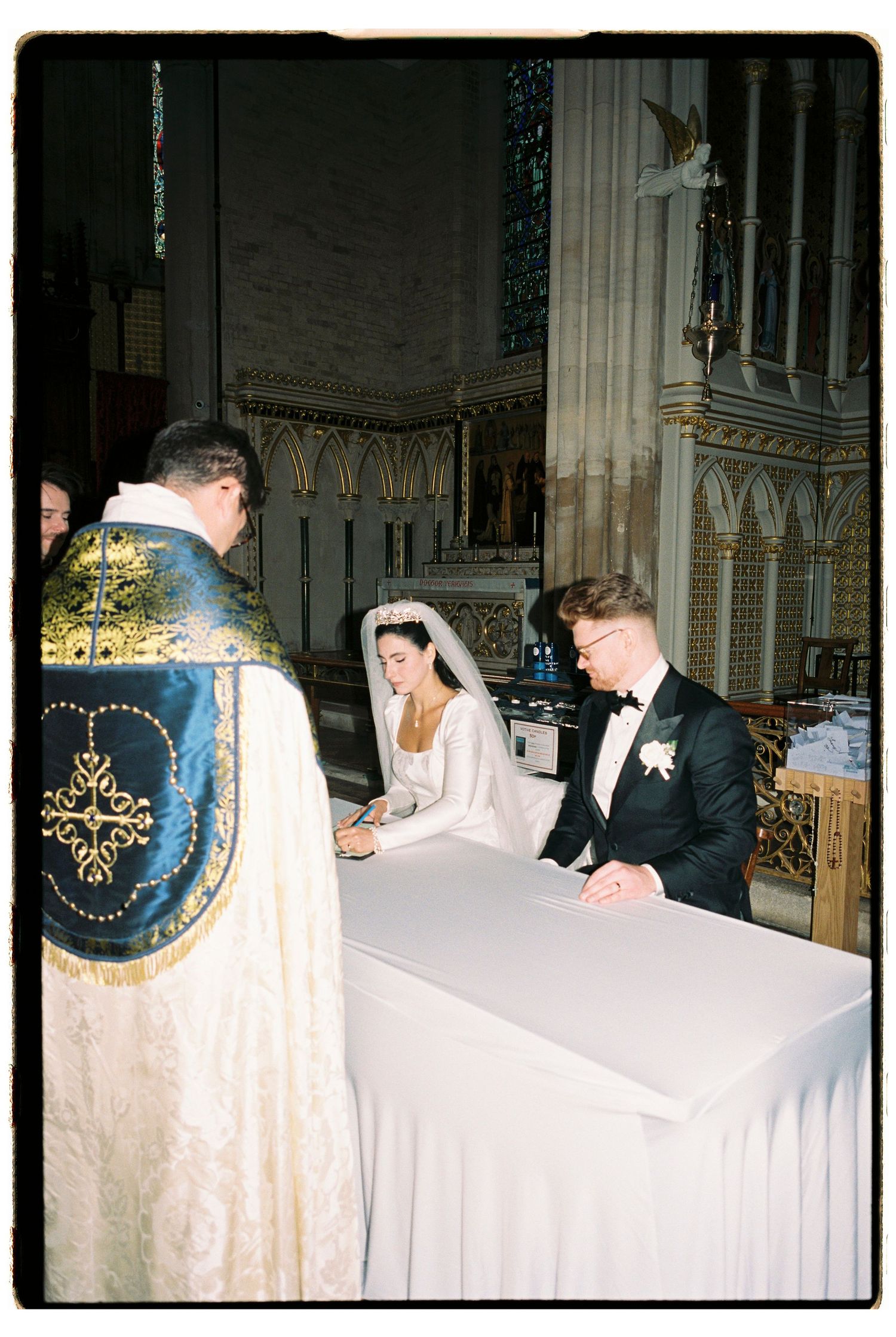Bride and groom signing wedding register at church altar with priest in ornate gold vestments.