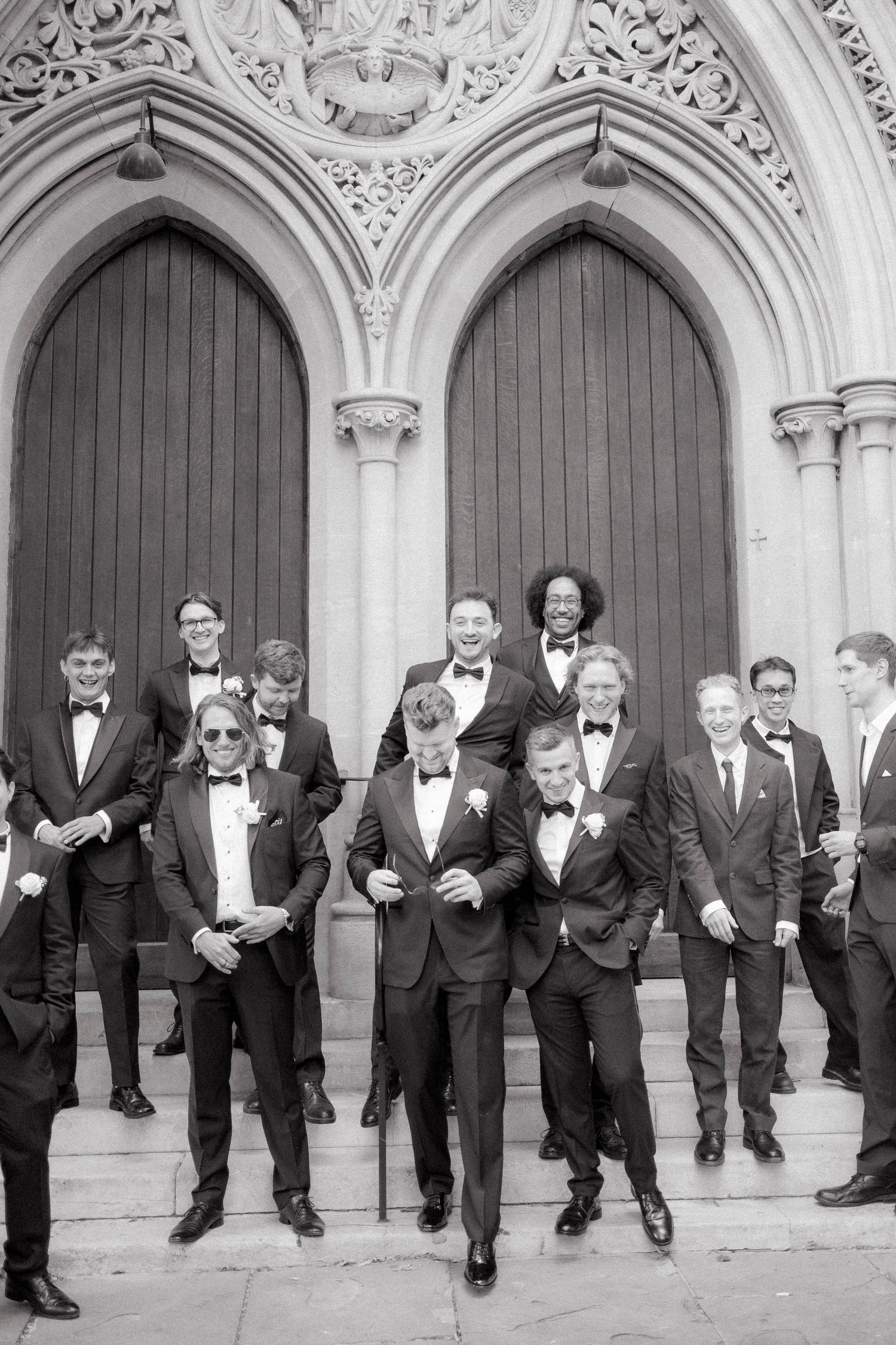 Groom and groomsmen in black tuxedos pose on church steps in a classic black and white wedding photo.
