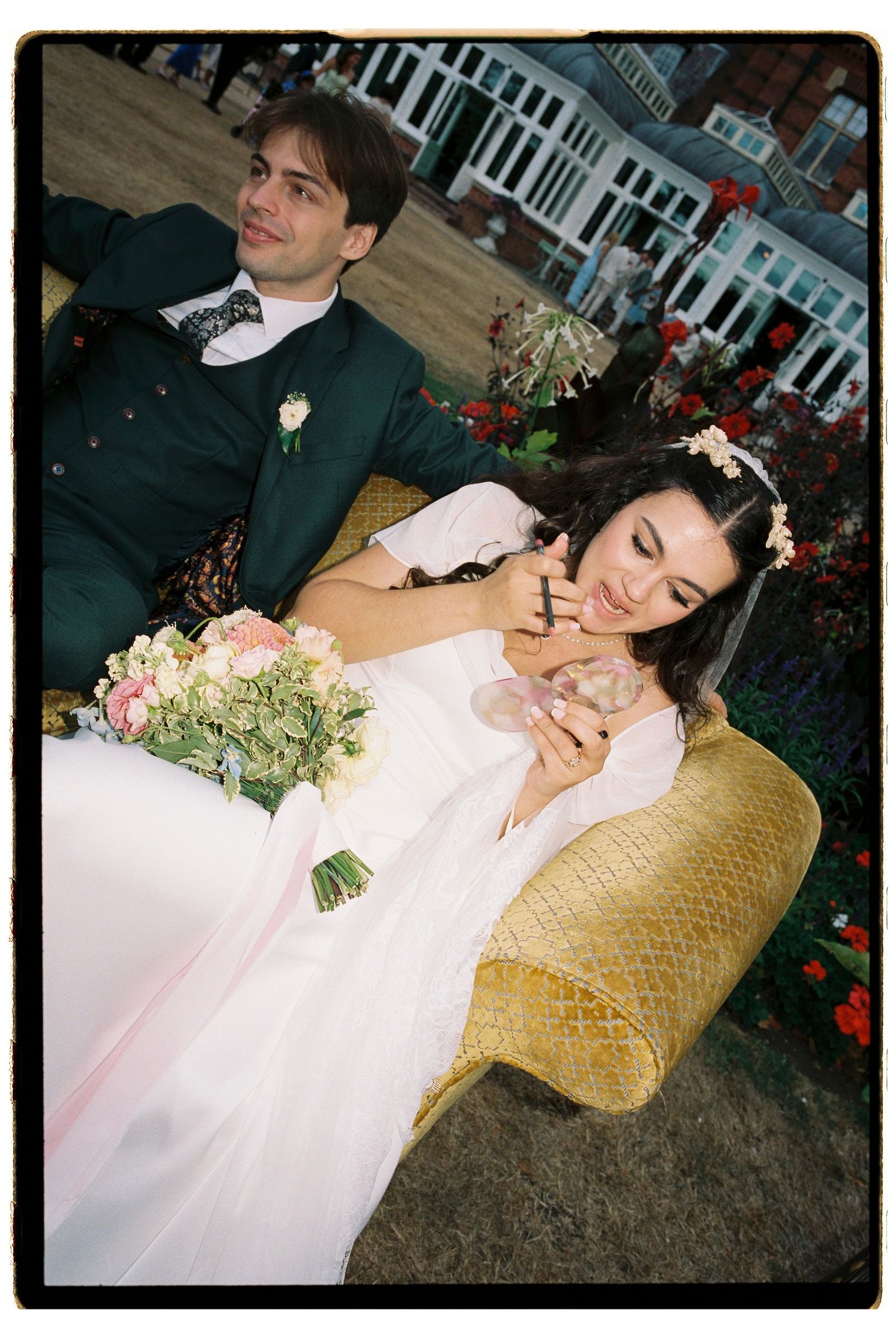 Bride in pink gown holds bouquet while groom in green suit sits beside her on golden chair at outdoor wedding.