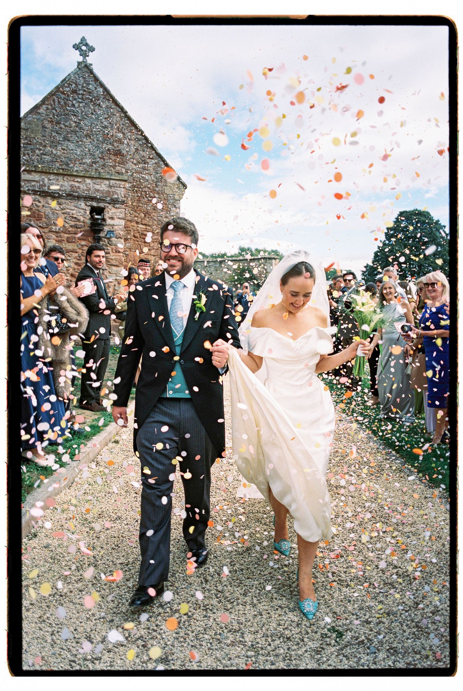 Newlyweds exit church in shower of colorful confetti surrounded by cheering guests.