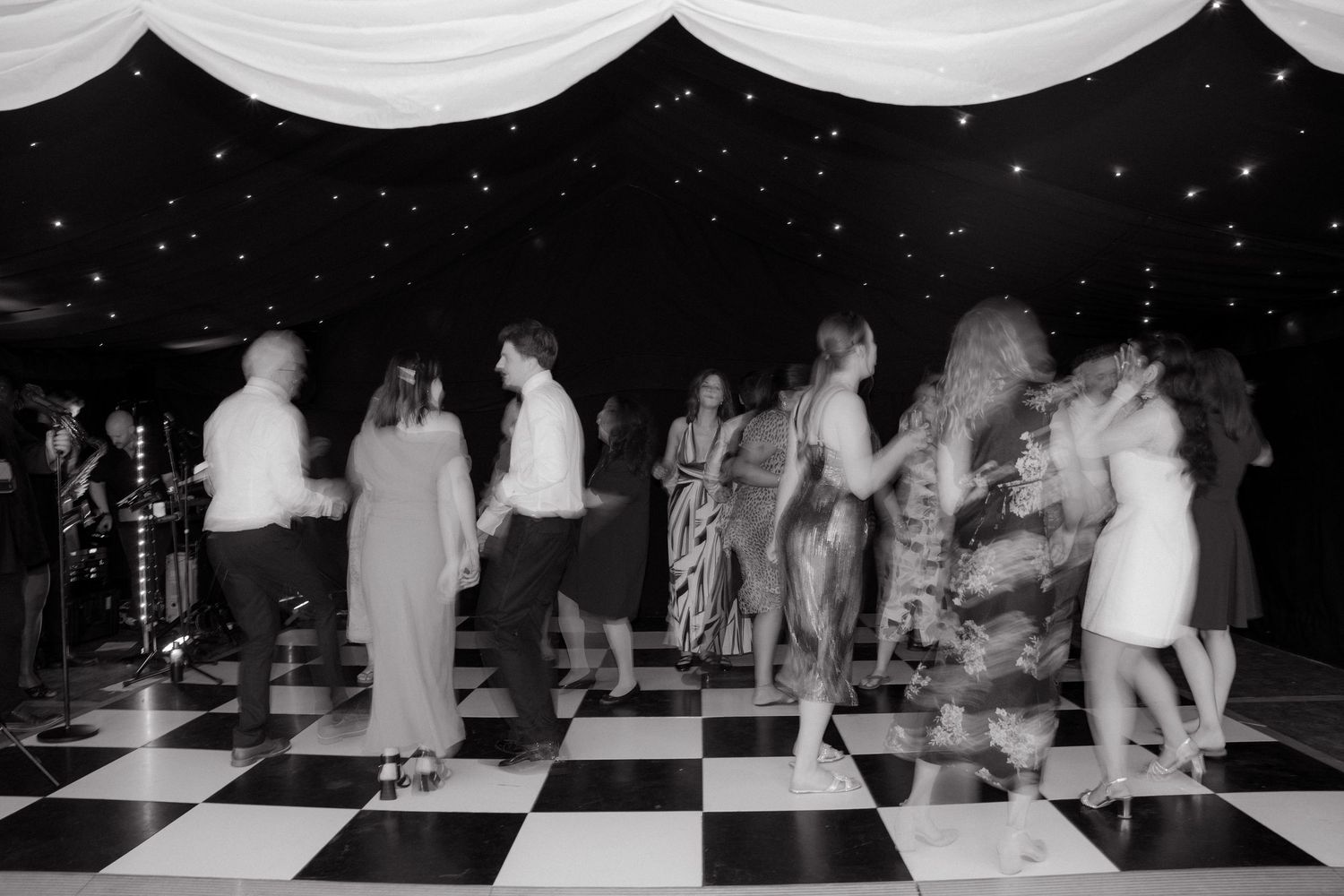 Wedding guests dancing on a black and white checkered dance floor under draped fabric ceiling.