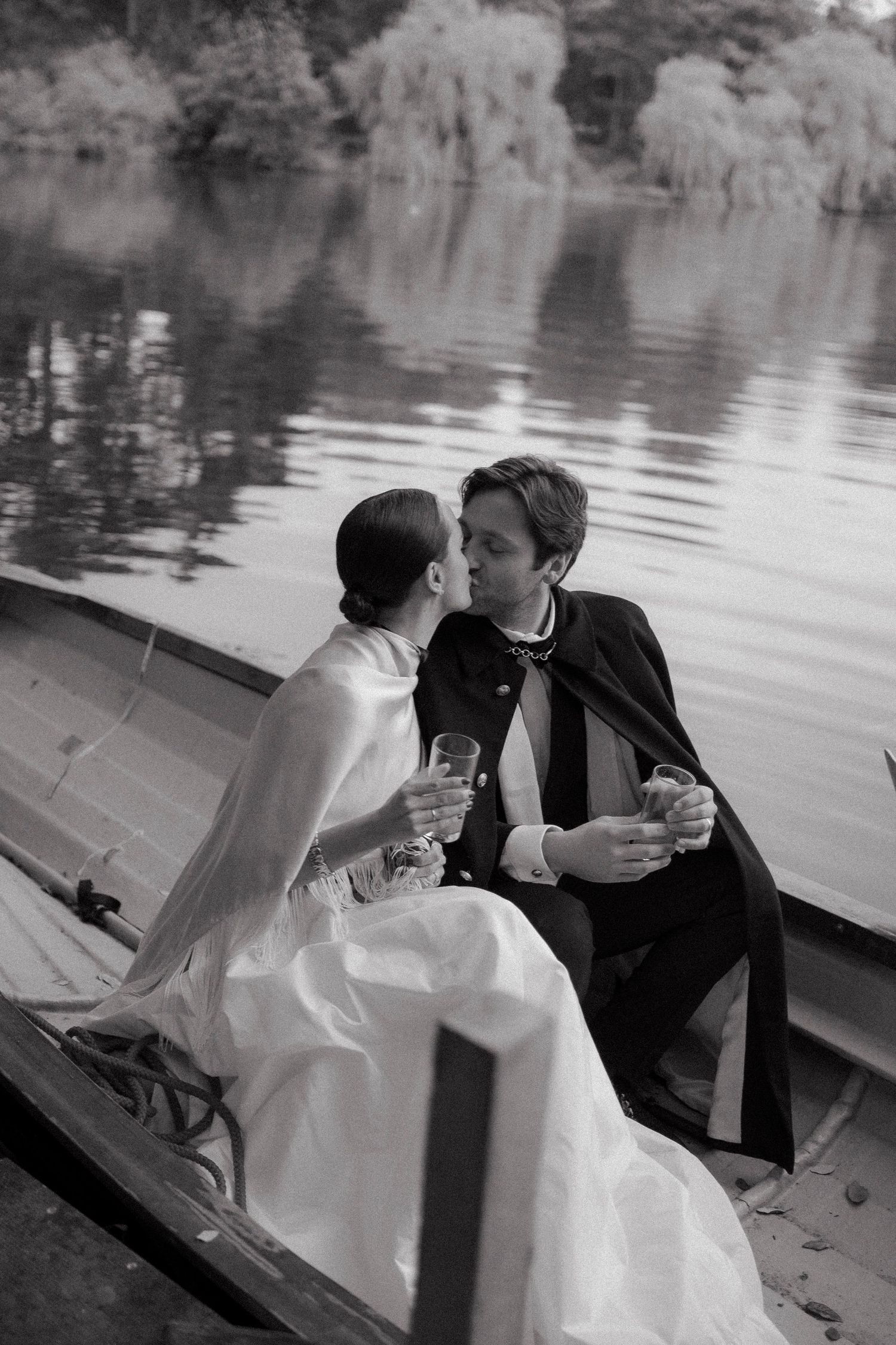 Black and white photo of a bride and groom kissing in a rowboat on a calm lake while holding champagne glasses.