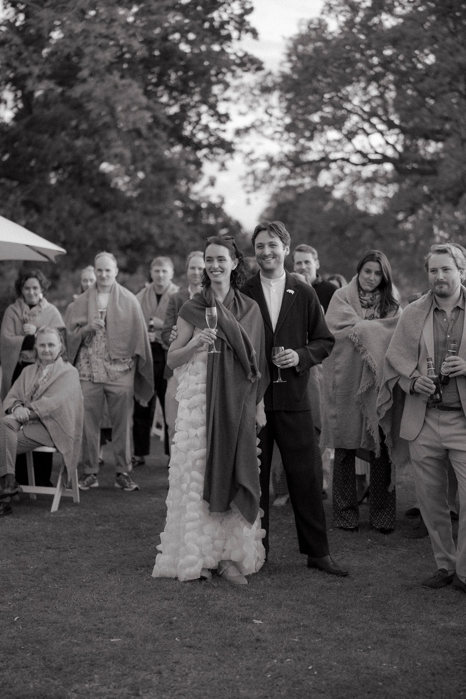 Bride in a ruffled white gown and groom in a dark suit hold champagne glasses and smile with wedding guests in blankets outdoors.