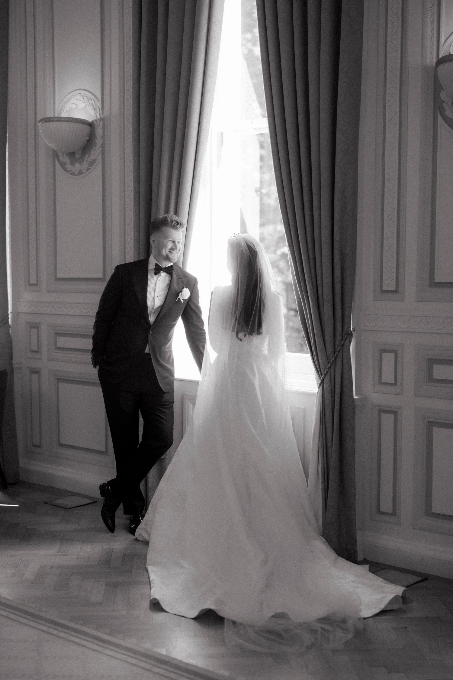 Black and white photo of bride and groom sharing intimate moment by tall window in elegant room.