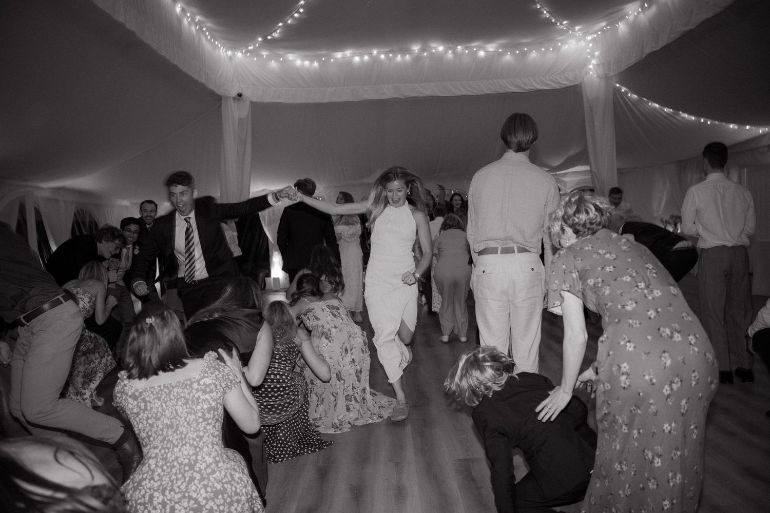 Black and white photo showing guests dancing and socializing at a wedding reception with string lights overhead.