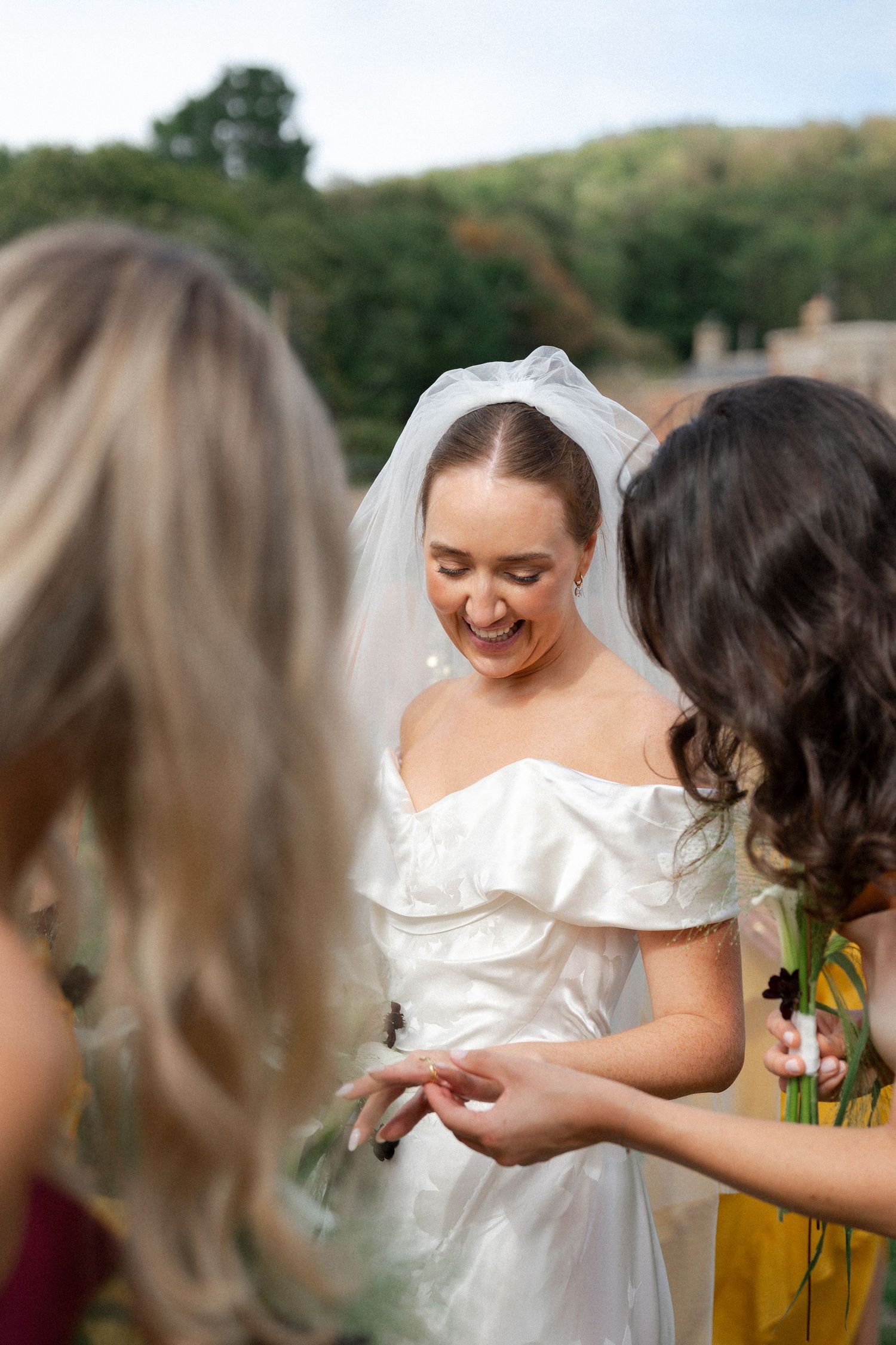 Smiling bride in white off-shoulder gown and veil admiring her ring with bridesmaids outdoors.