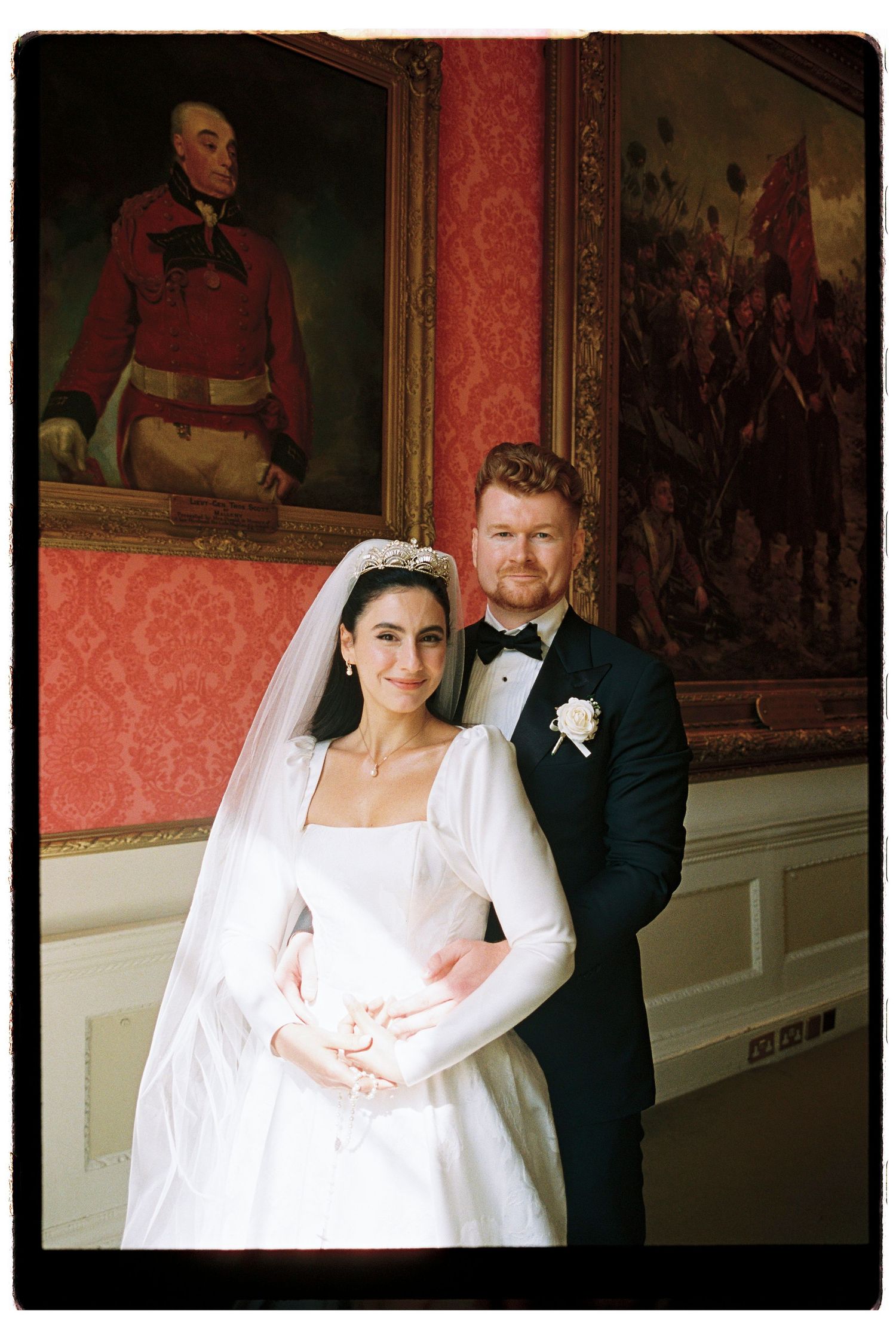 Bride in tiara and groom in tuxedo pose before historic military portrait in elegant red room.