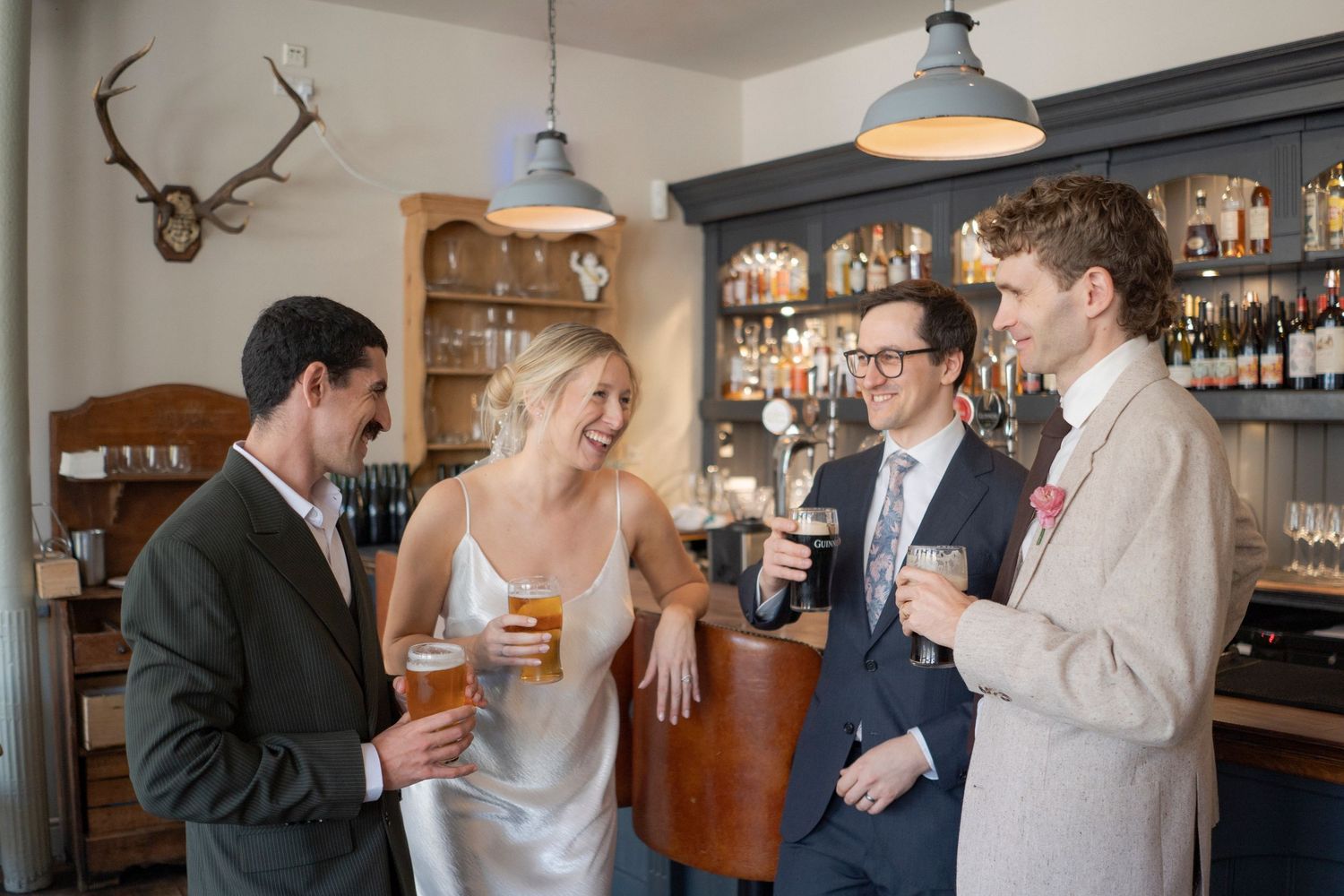 Four elegantly dressed guests laughing and enjoying drinks together at a stylish bar during a wedding reception.