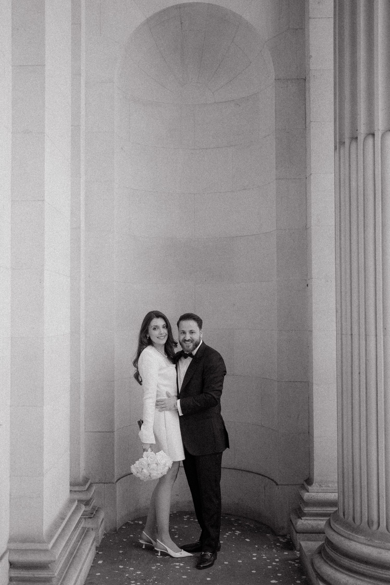 Black and white photo of a couple embracing between classical stone columns, woman holding white flowers, both dressed formally.
