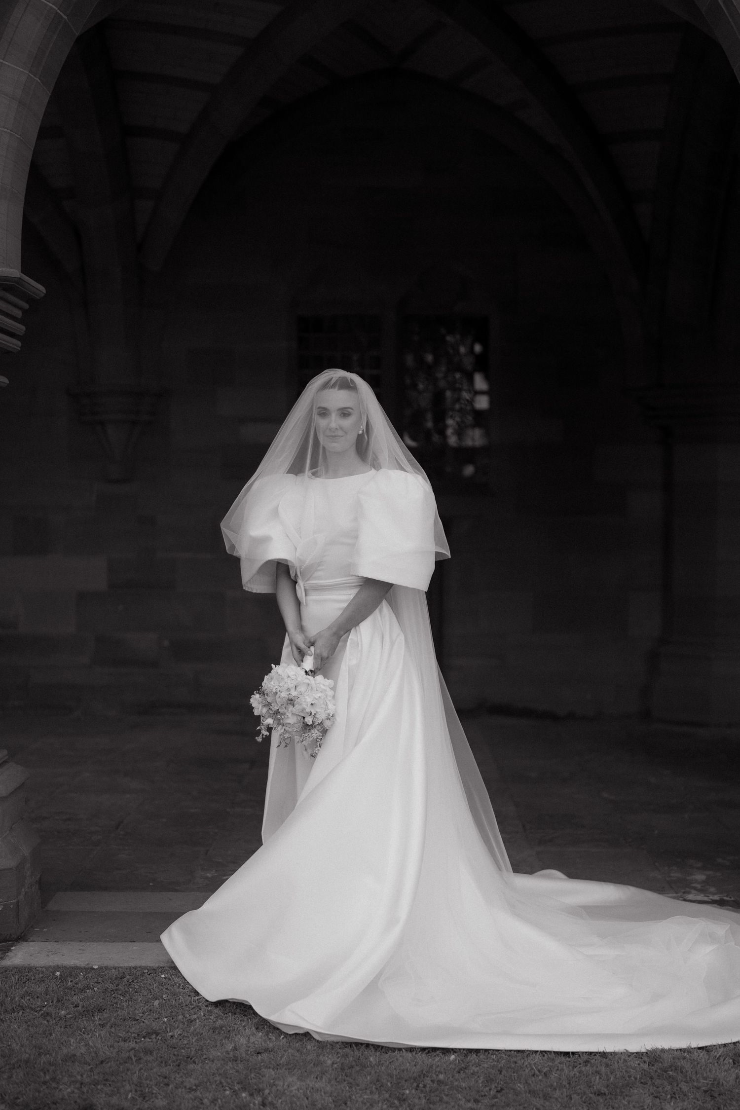 Black and white portrait of a bride in a flowing dress with statement sleeves and long veil outdoors.