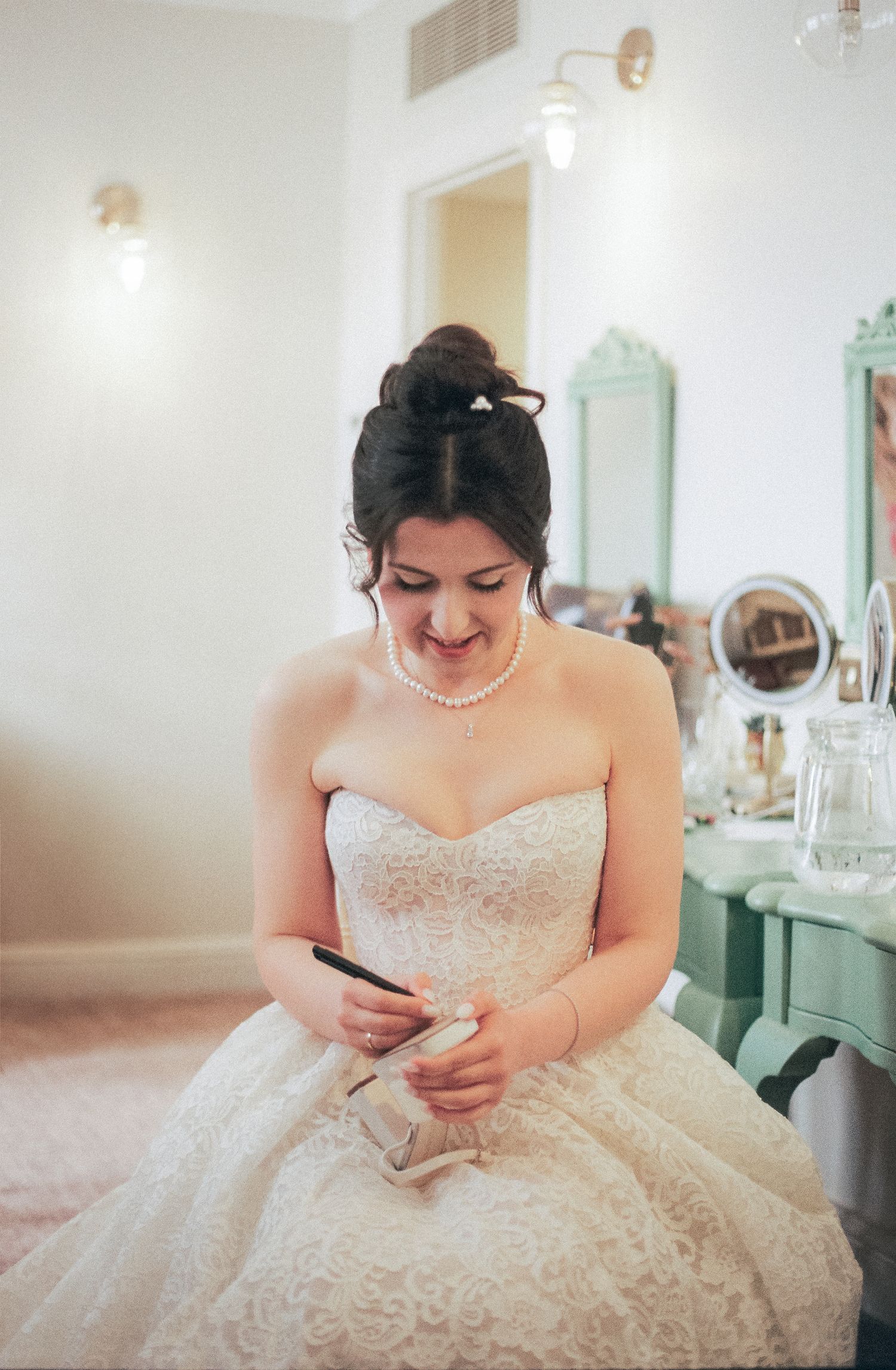 Bride in an elegant lace wedding gown checks her phone while sitting in a dressing room.