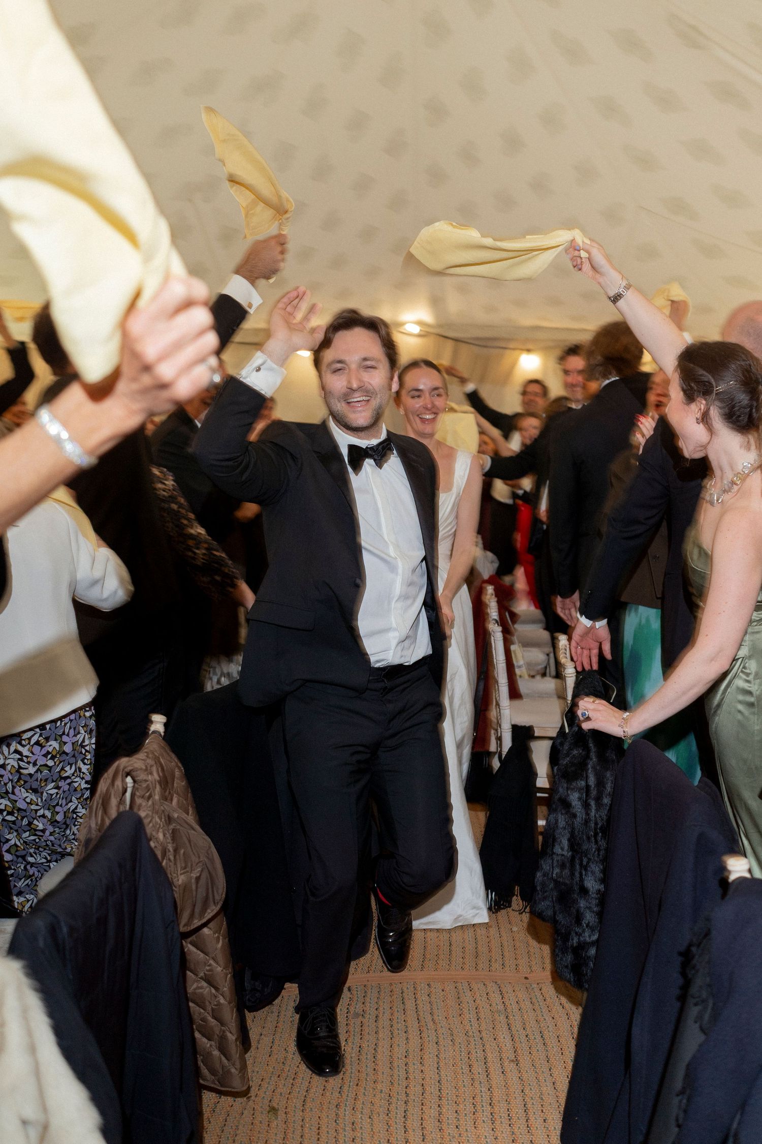 Smiling groom in a black bow tie dances down the aisle as wedding guests wave white napkins in the air at the reception.