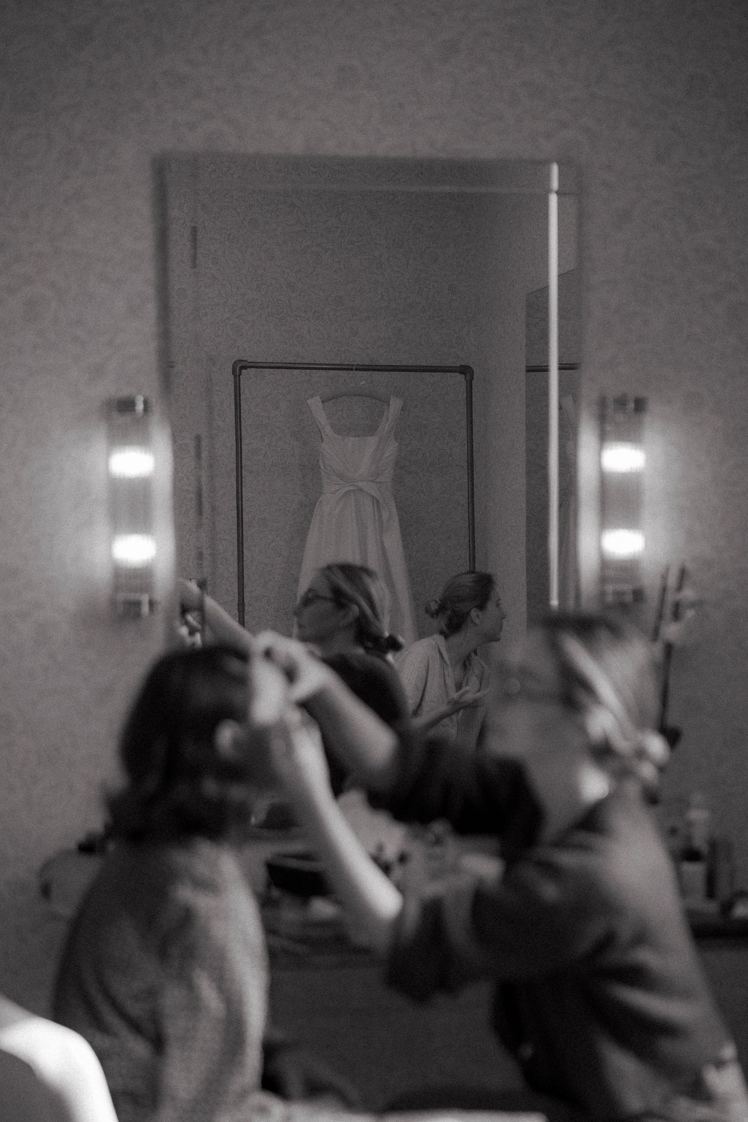 Black and white photo of a bride getting ready as two women help with her hair, a wedding dress reflected in the mirror.